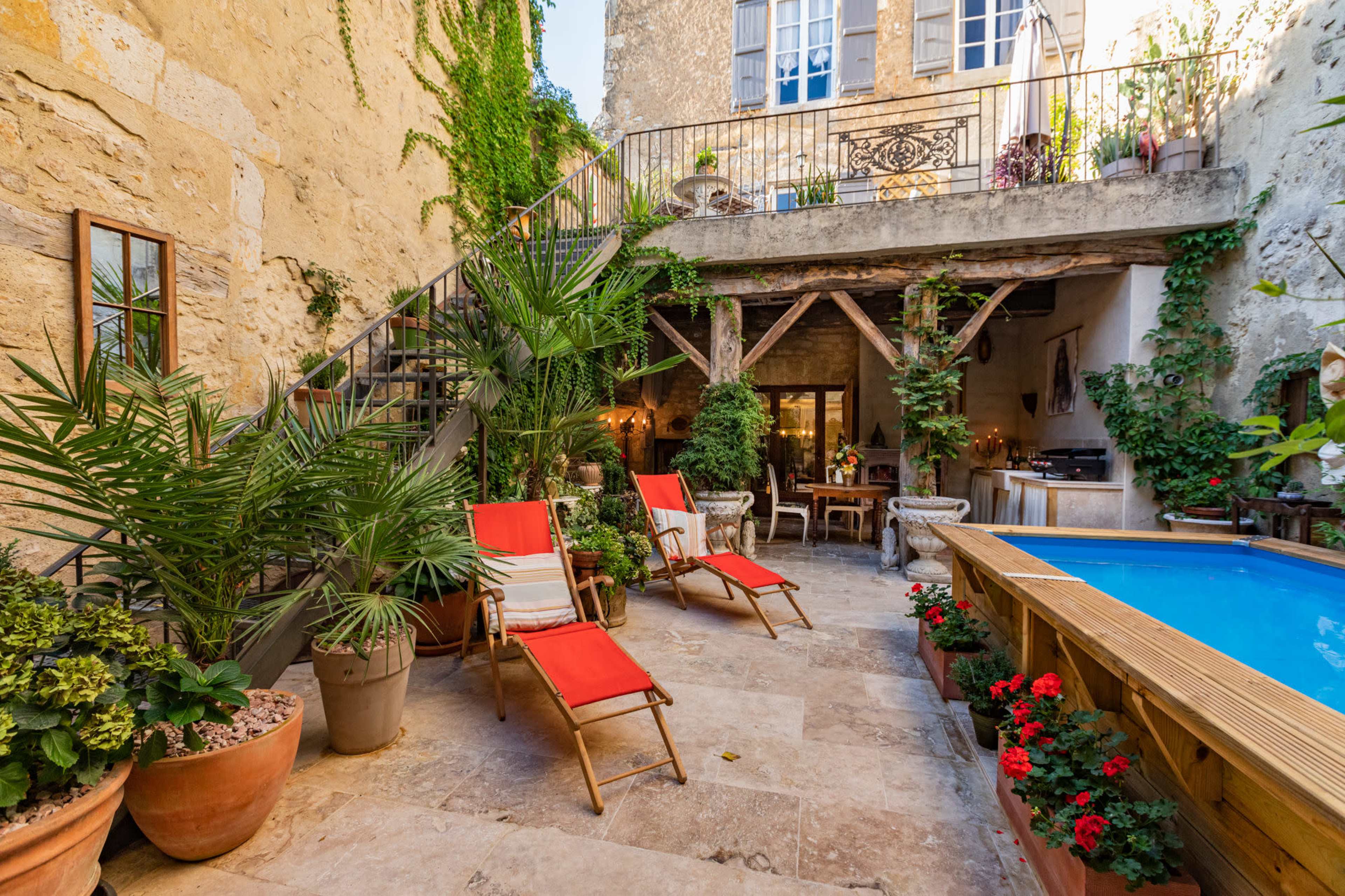 A courtyard features a small blue pool surrounded by potted plants, red lounge chairs, and an adjacent dining area under a wooden structure.