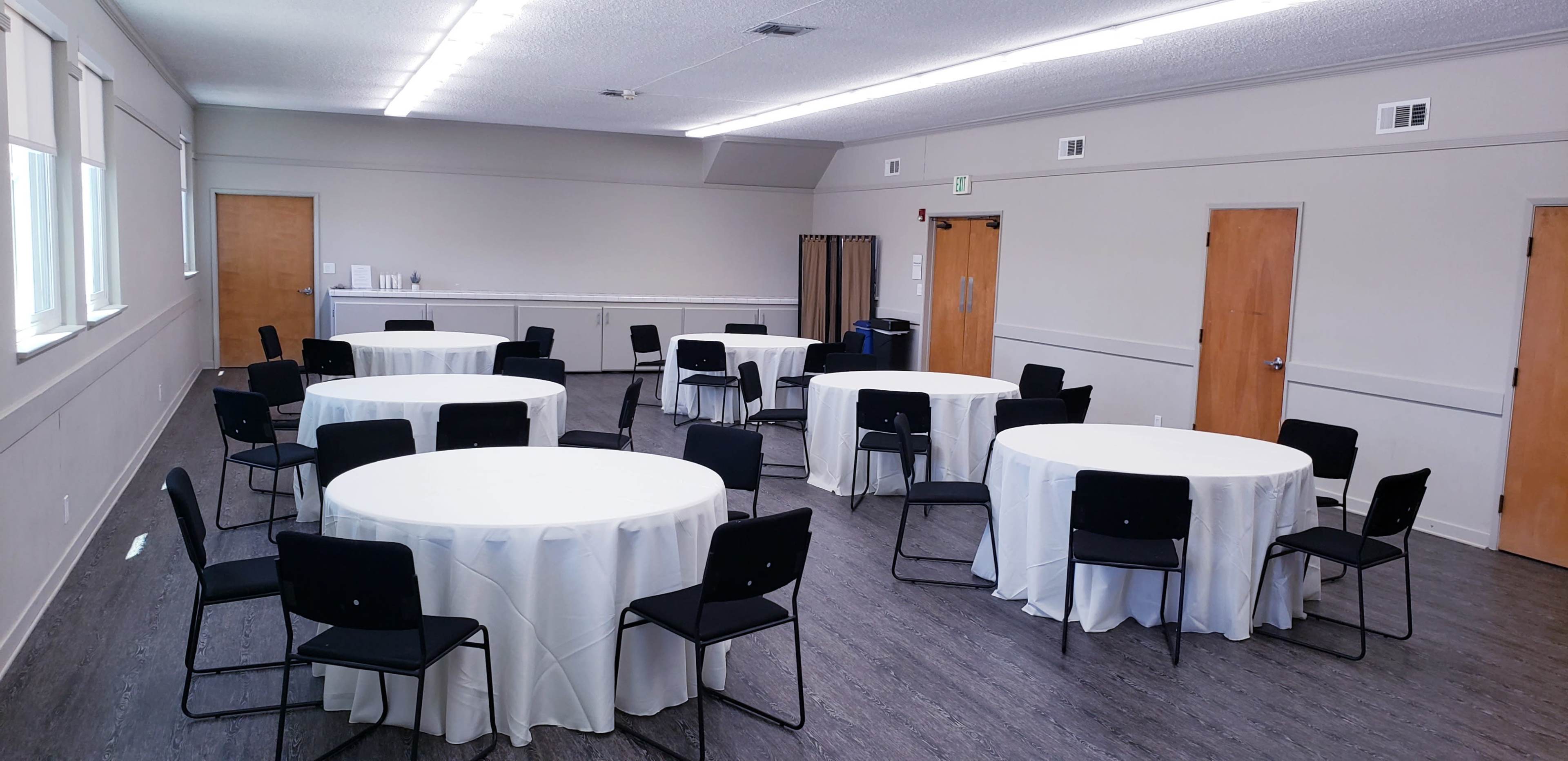 A meeting room with eight round tables covered in white tablecloths, each surrounded by black chairs.