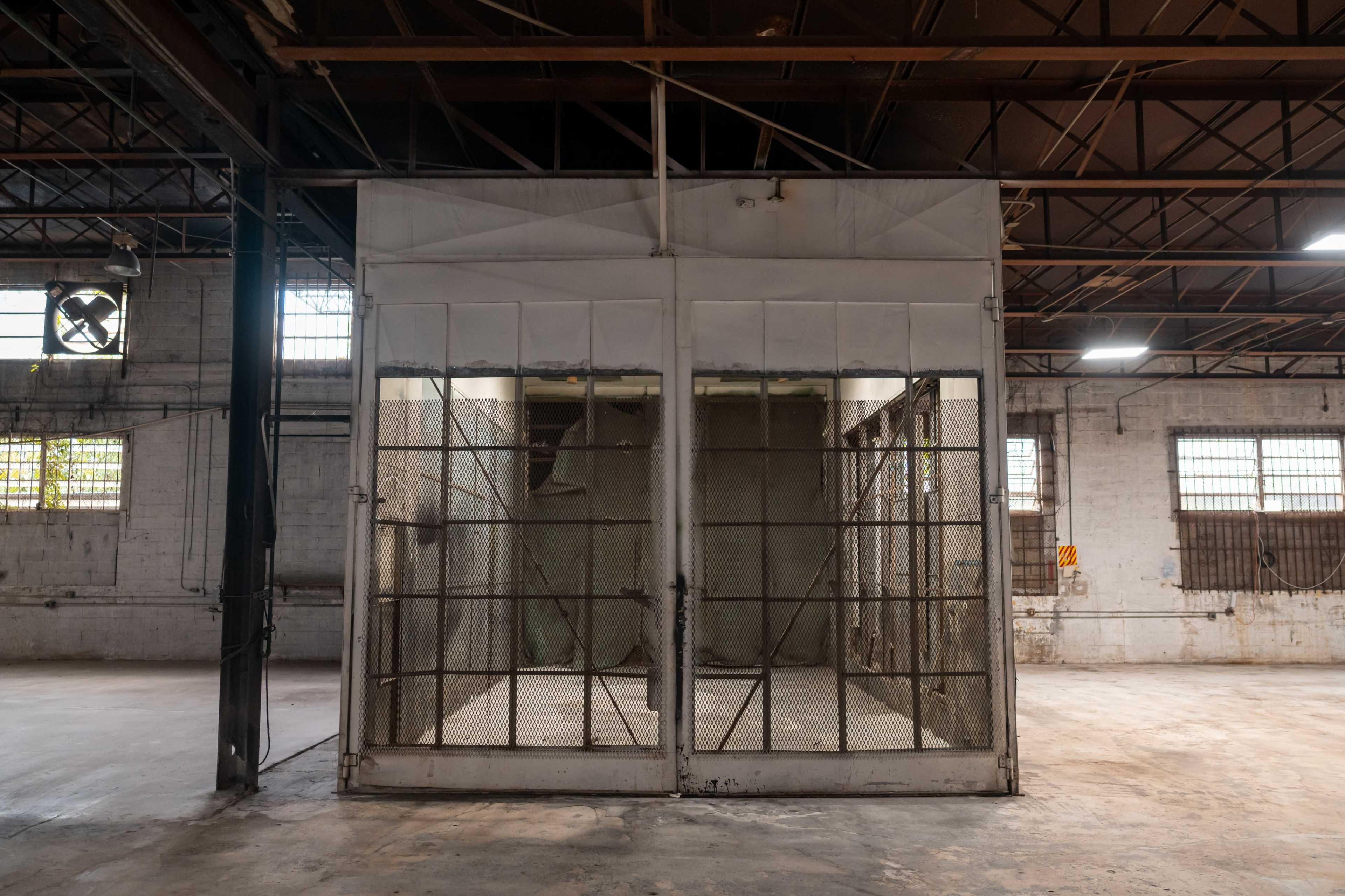 The image shows a large, empty industrial space featuring a fenced-in enclosure made of metal and mesh.