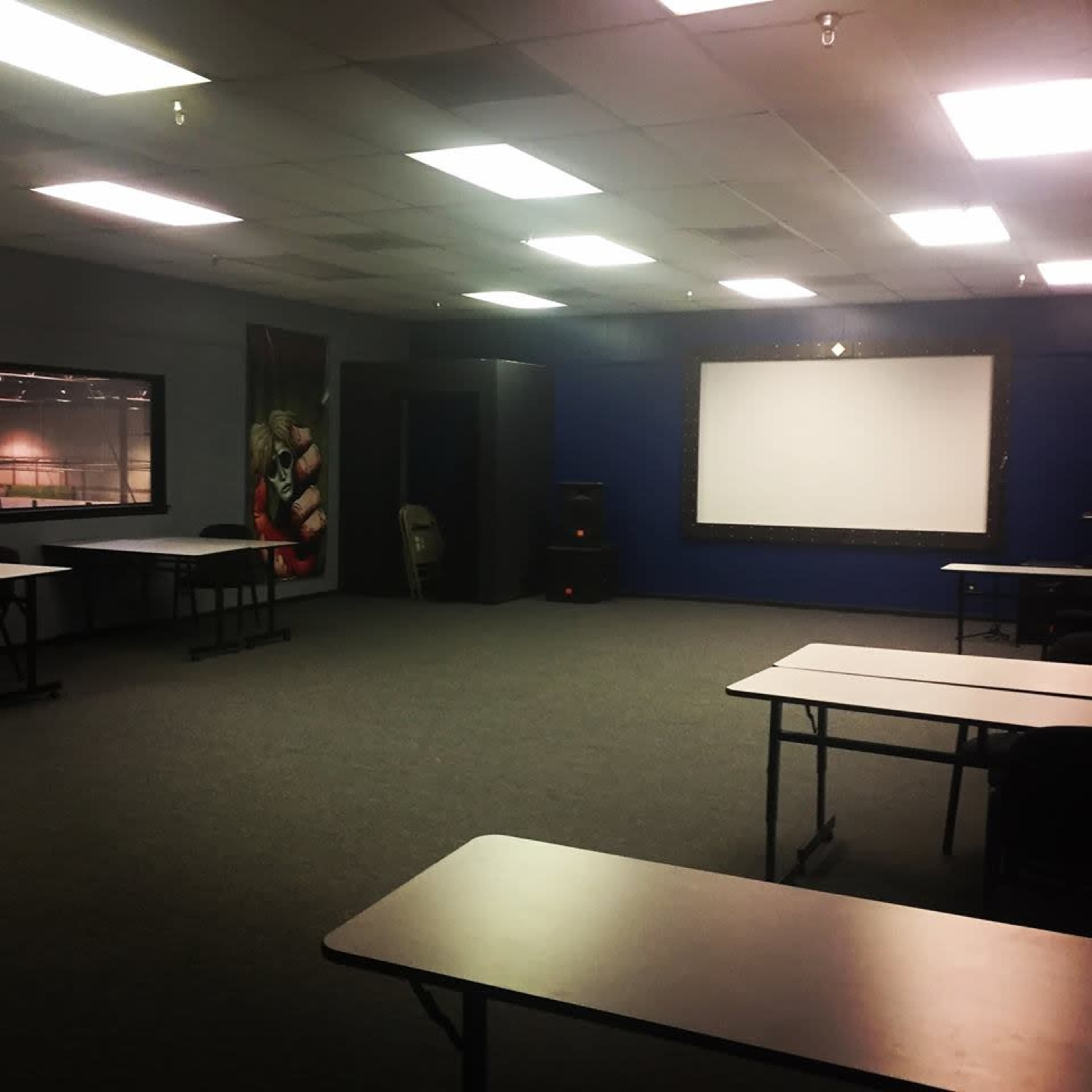 The image shows a spacious, empty room with several tables arranged facing a blank projector screen on a blue wall.