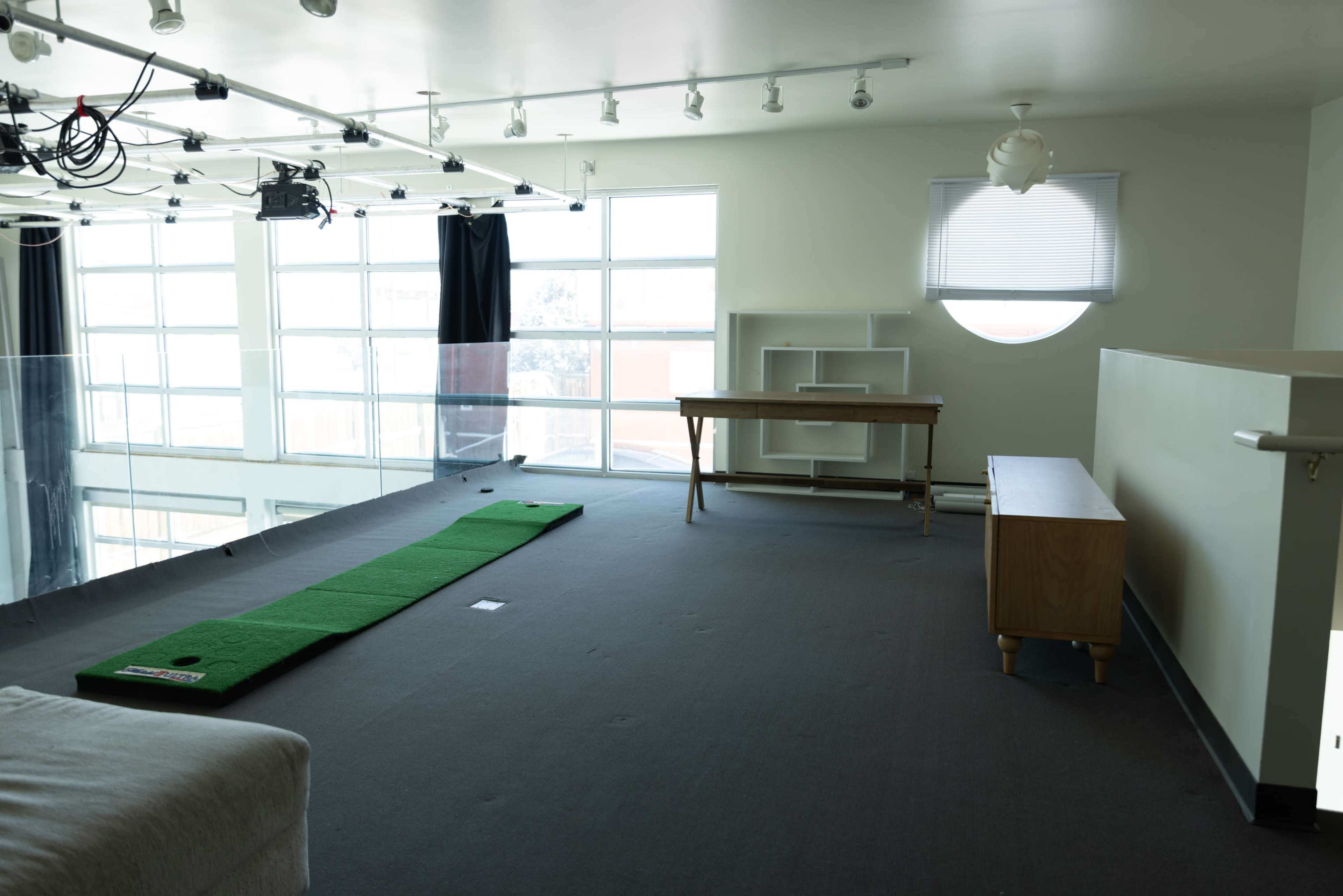 The image shows a spacious, brightly lit room with large windows, featuring a putting green on the floor, a wooden table, and a sideboard.