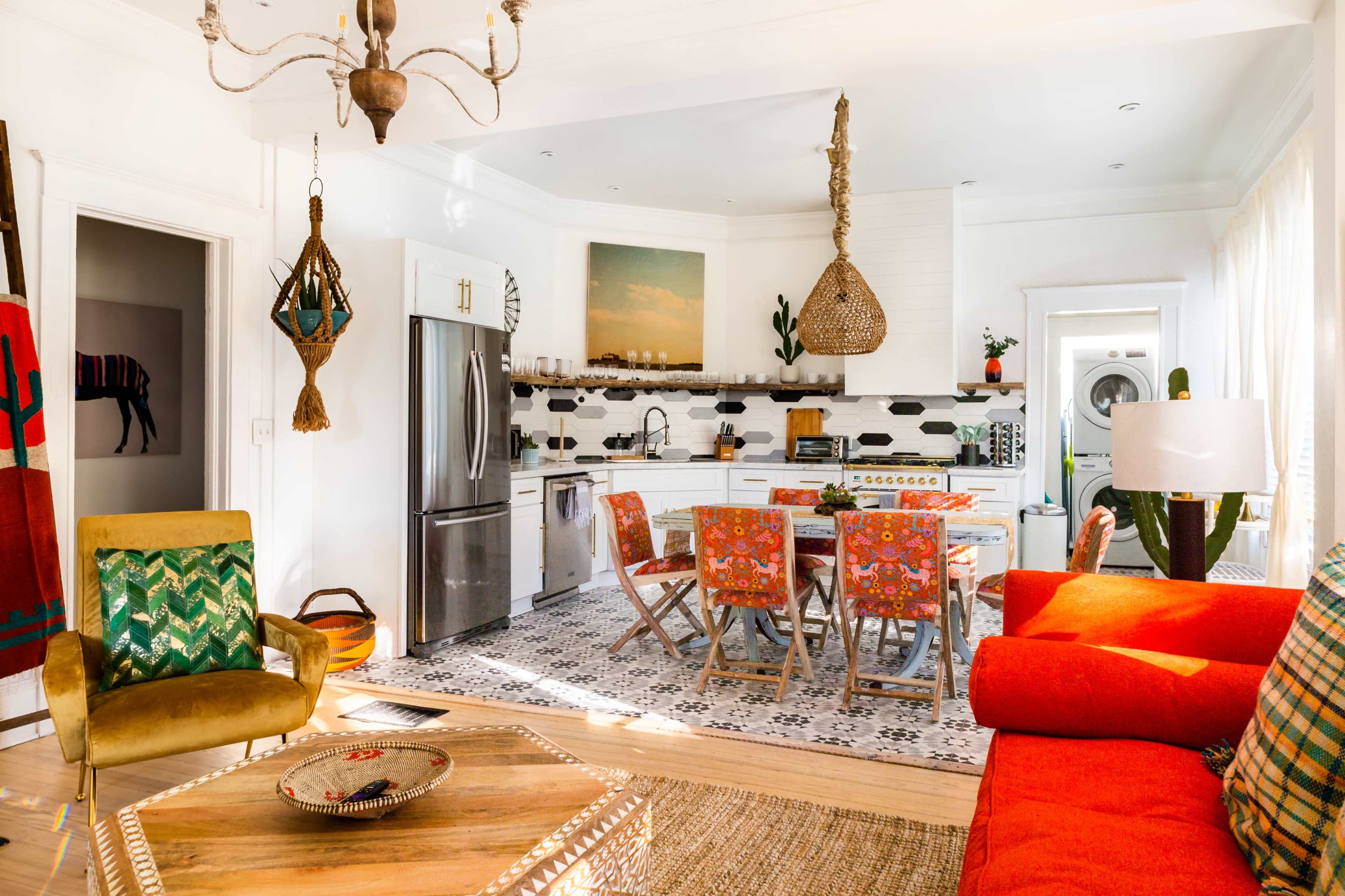 A bright, colorful kitchen and living area feature a dining table surrounded by patterned chairs, with a modern refrigerator and decorative elements like plants and rugs.