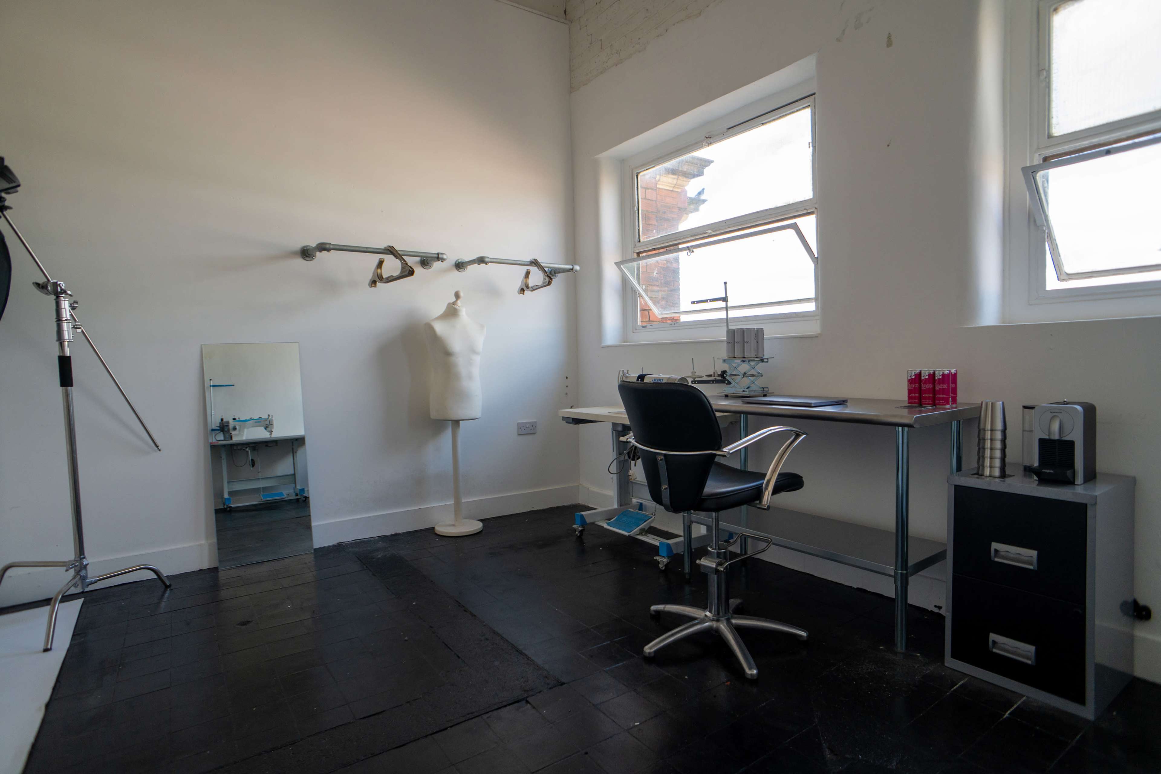 The image shows a minimalist studio space with a mannequin, a desk with a chair, a mirror, and a small black filing cabinet.