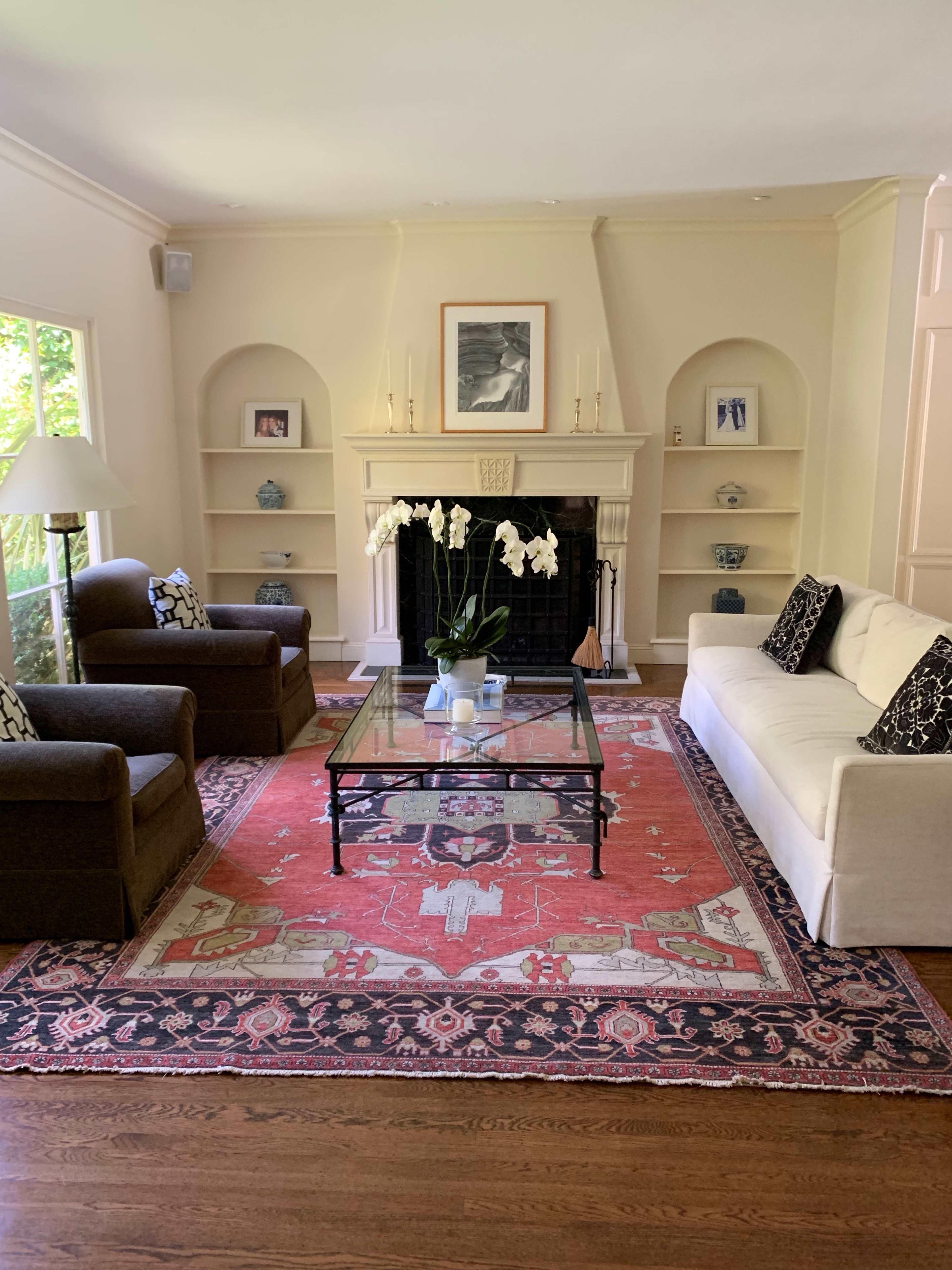 The living room features two armchairs and a white sofa arranged around a glass coffee table on a red patterned rug, with a fireplace and built-in shelves in the background.