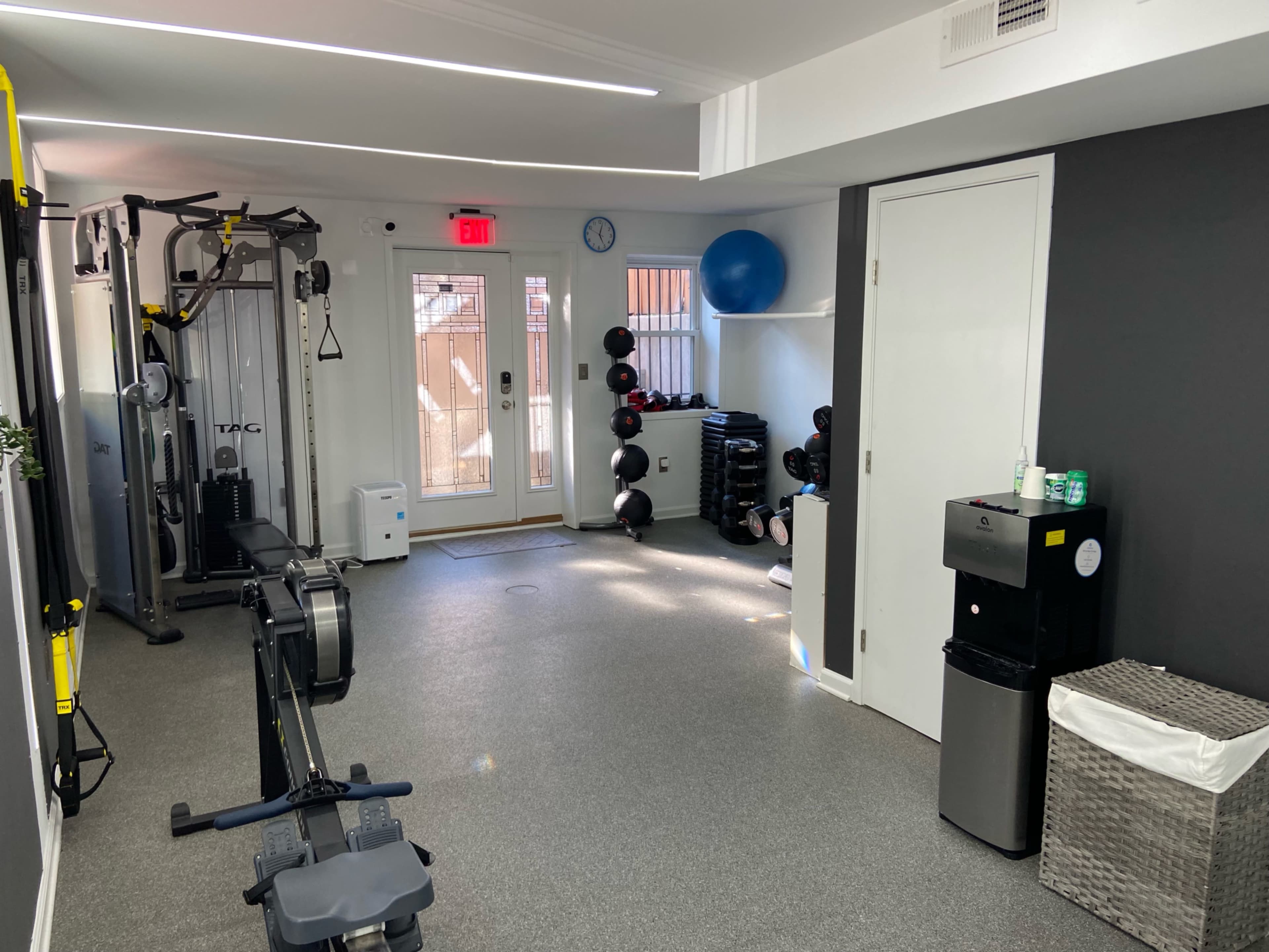 A well-lit home gym equipped with exercise machines, weights, a blue stability ball, and a water cooler, with a door leading to an outdoor area.