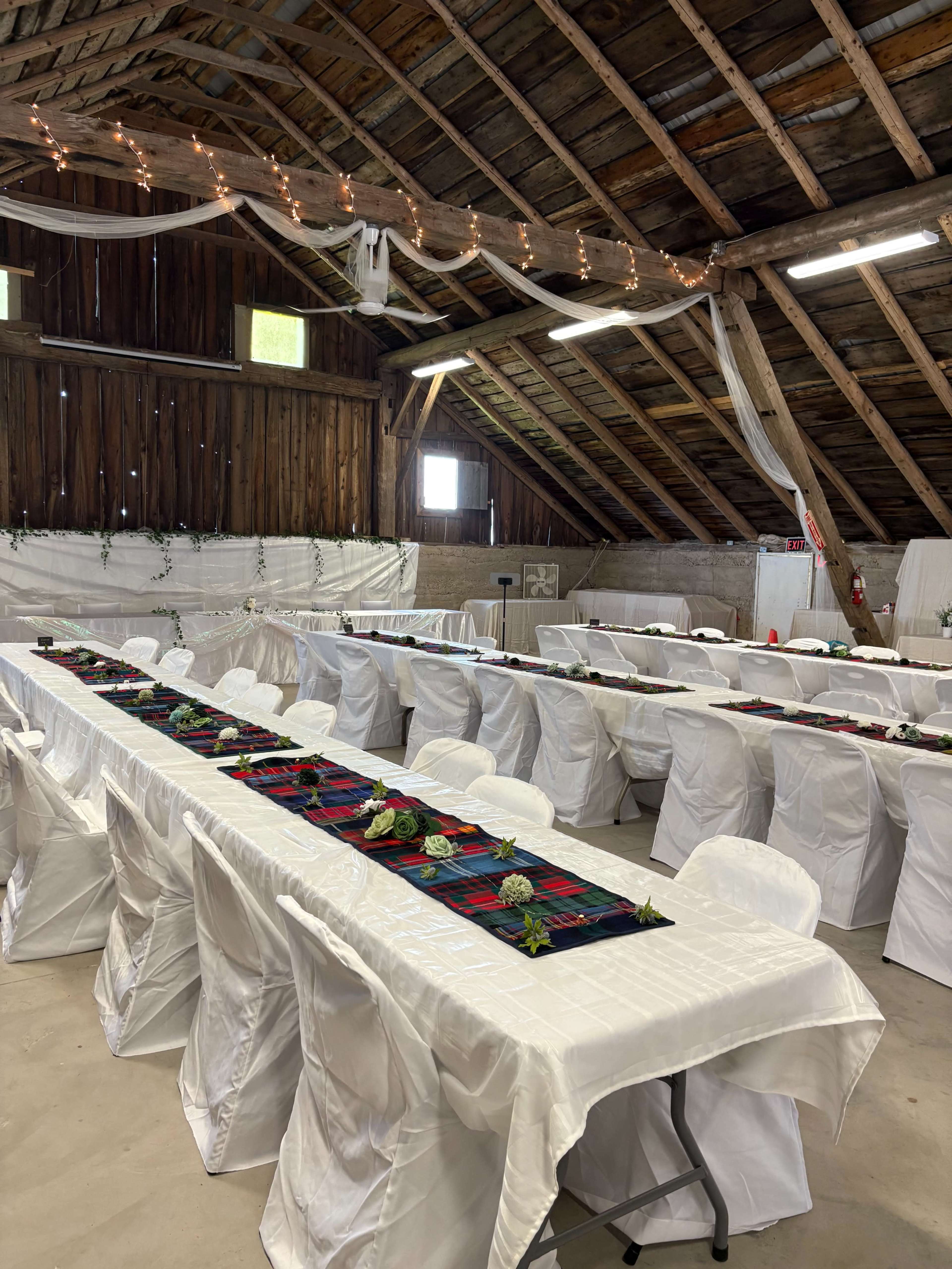 A spacious barn interior features several long tables covered with white tablecloths, decorated with plaid runners and green floral arrangements.