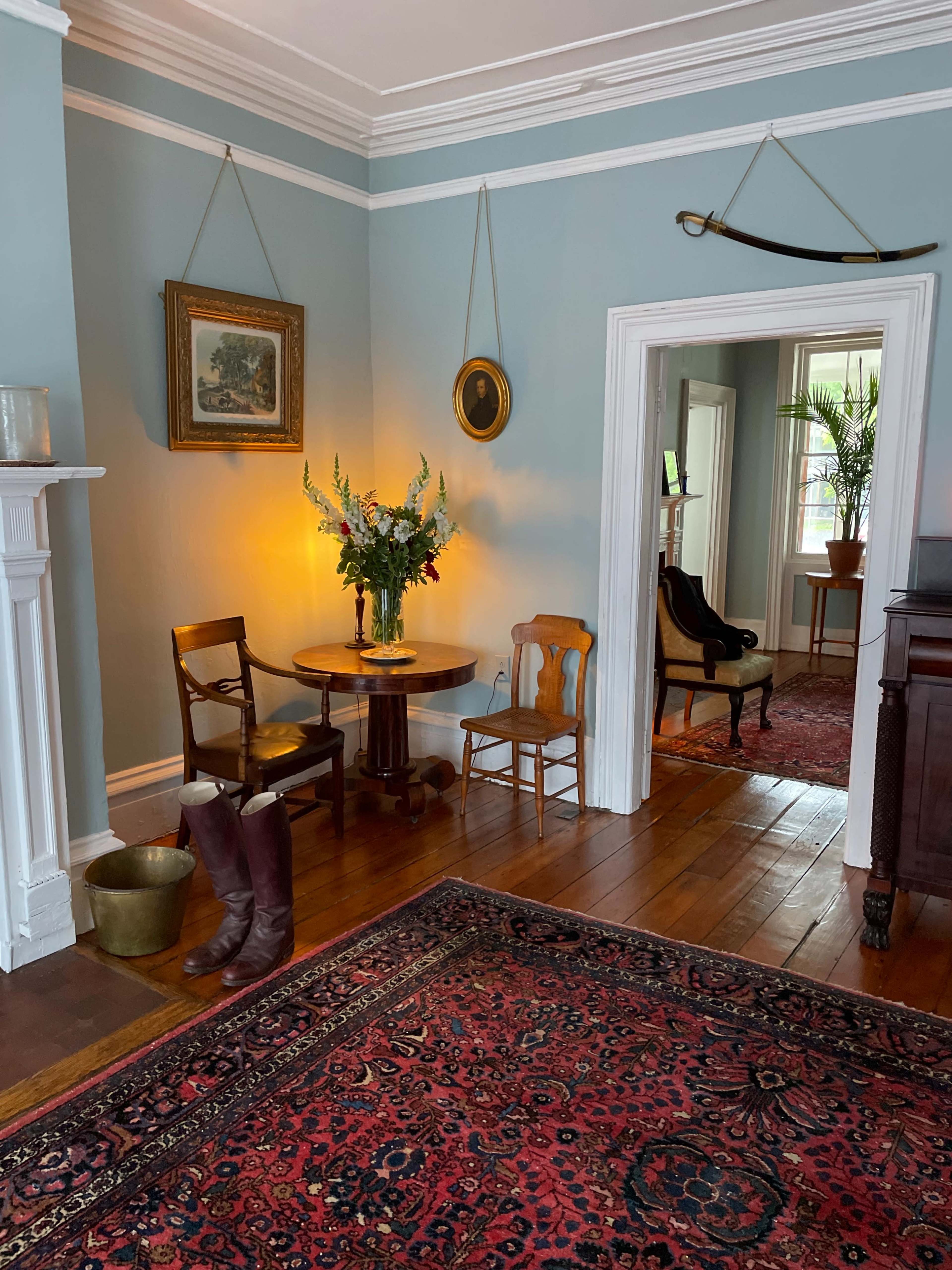 The image shows a cozy room with a round table and chairs, a floral arrangement on the table, and a view into an adjacent room through an open doorway.