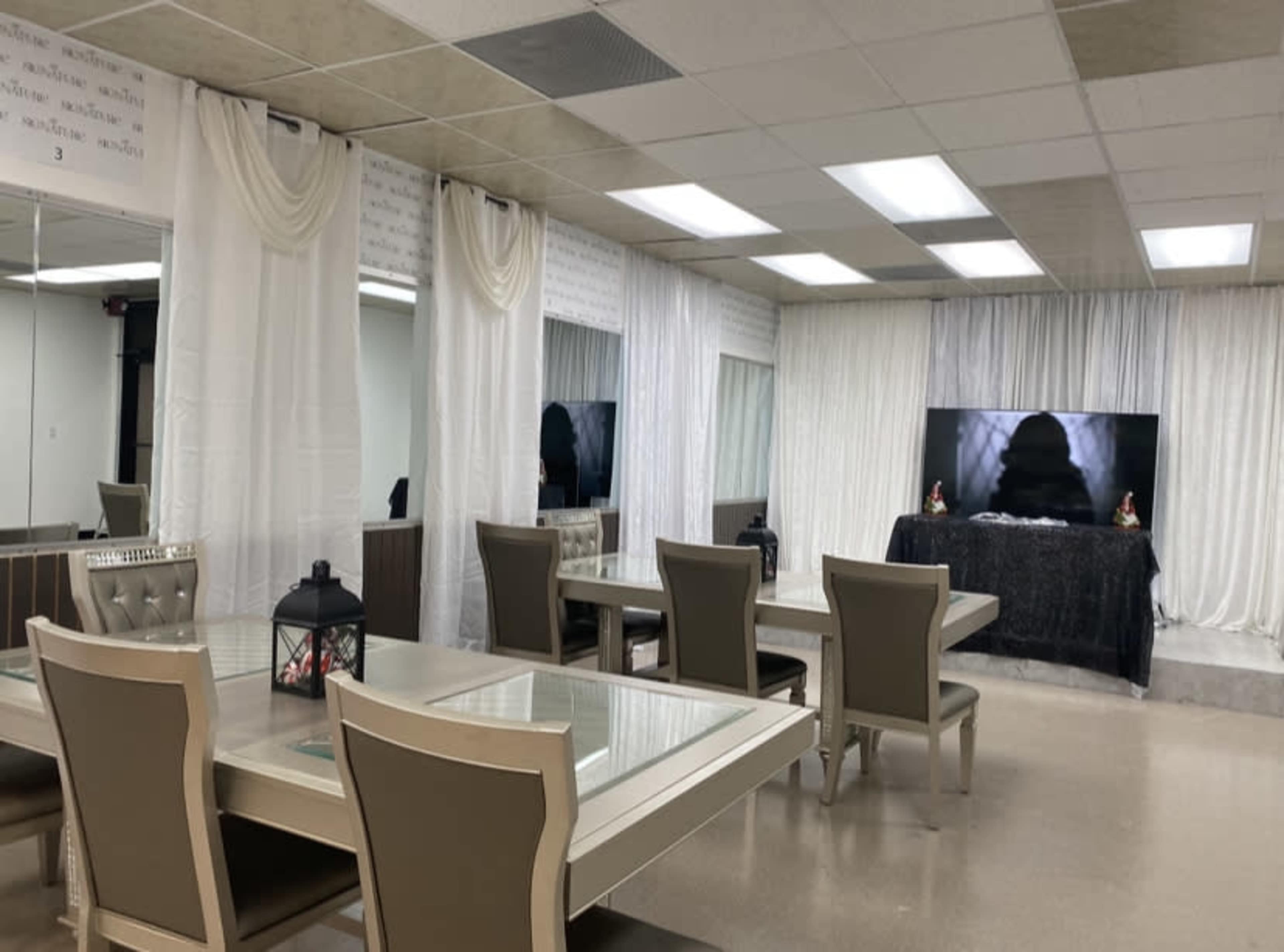 The image shows a spacious, brightly lit room with multiple tables and chairs, draped white curtains, and a television screen at one end.