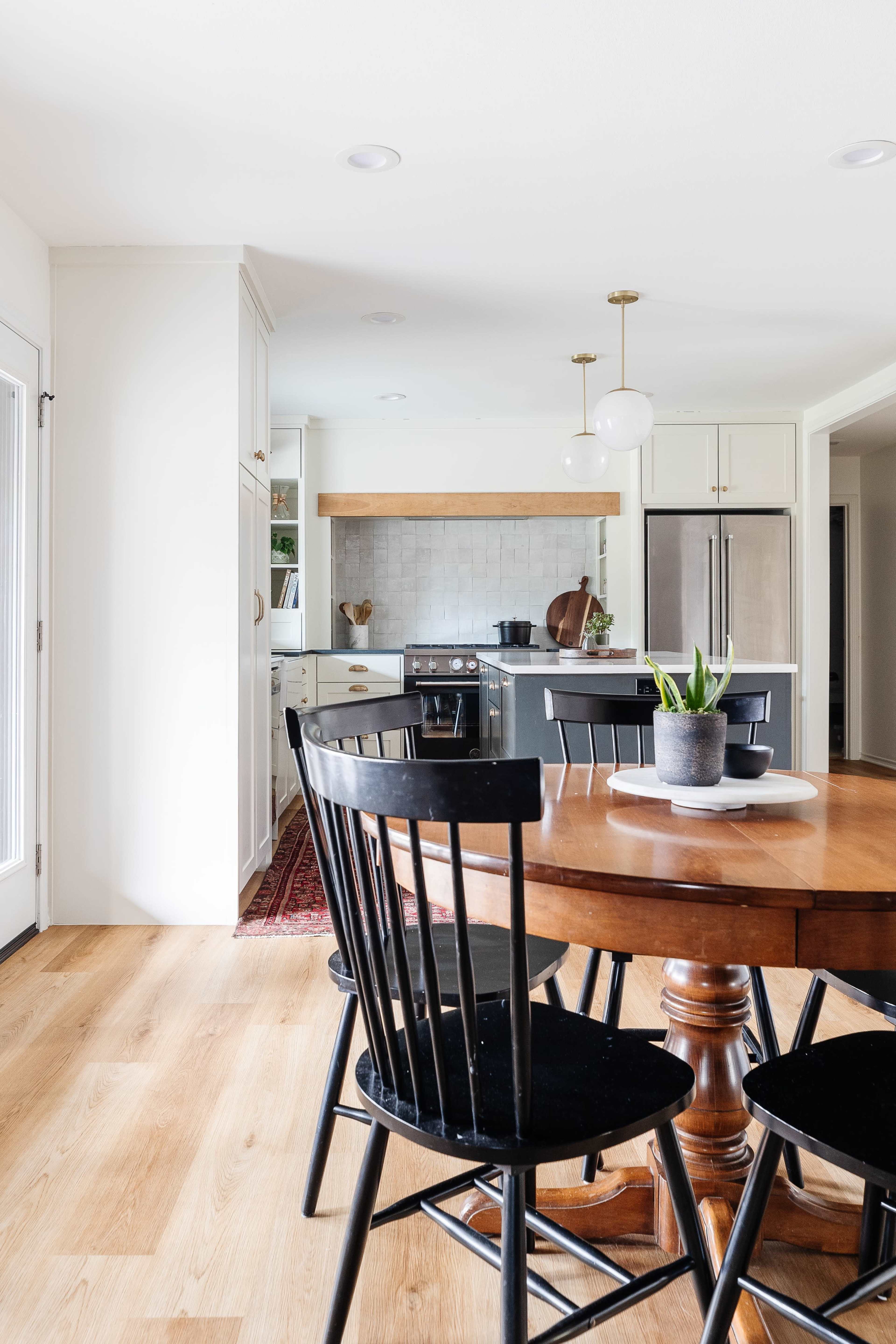 A wooden dining table with black chairs sits in a bright kitchen featuring light cabinets and a gray island.