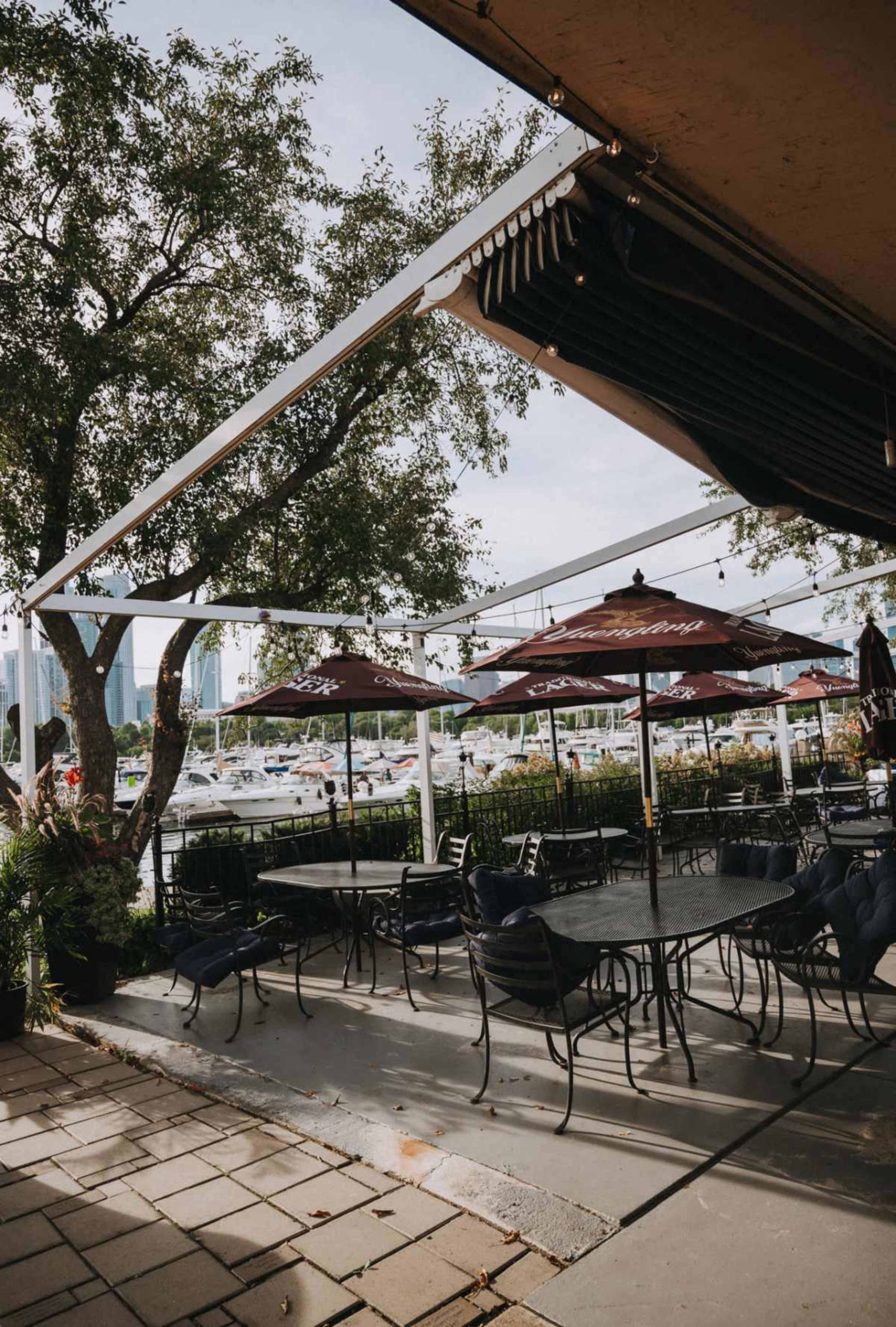 The image shows a patio area of a restaurant with metal tables and chairs, umbrellas, and a view of boats docked nearby.