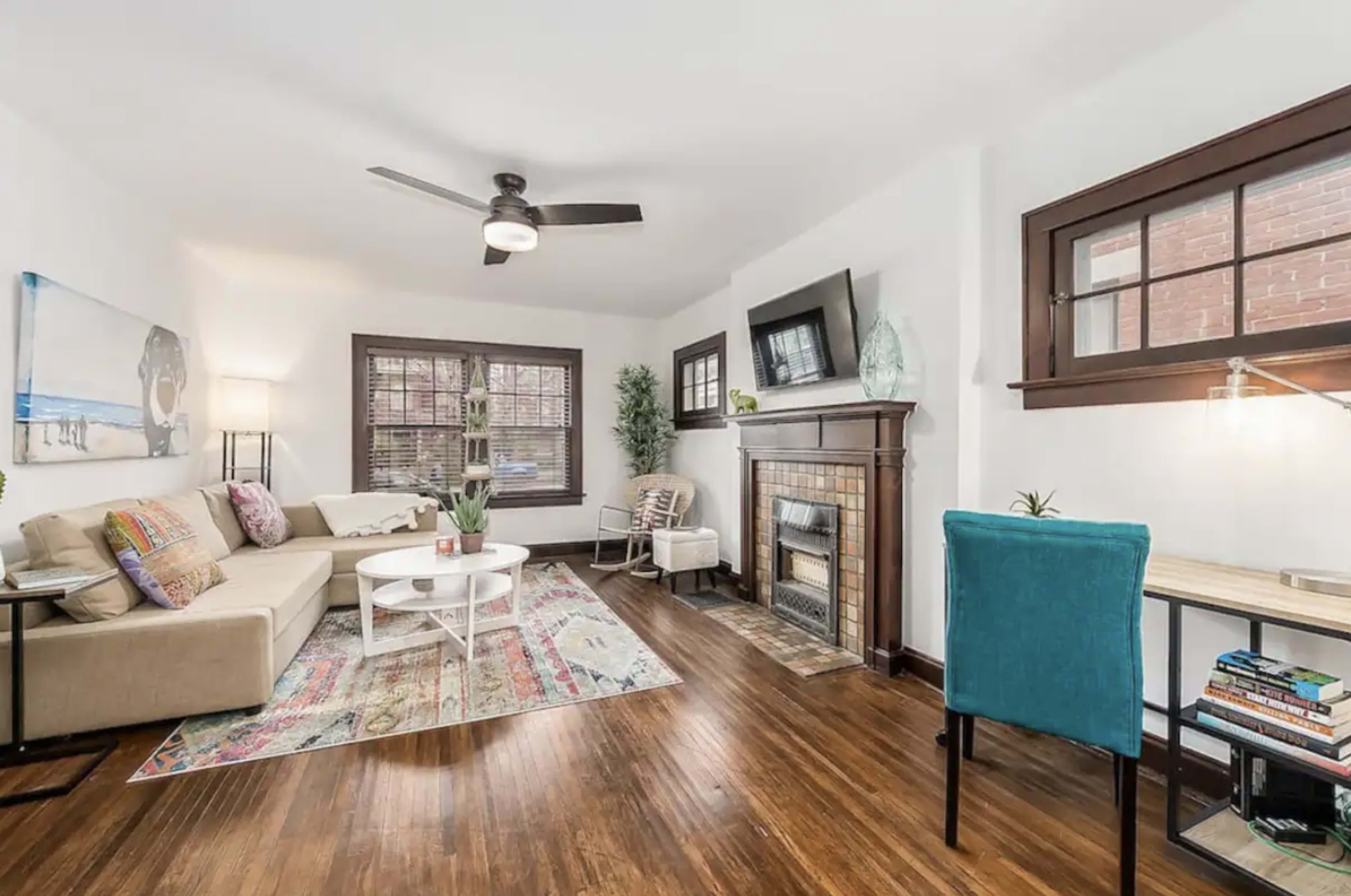 The living room features a beige couch, a white coffee table, a fireplace with a TV above it, and a workspace with a blue chair.
