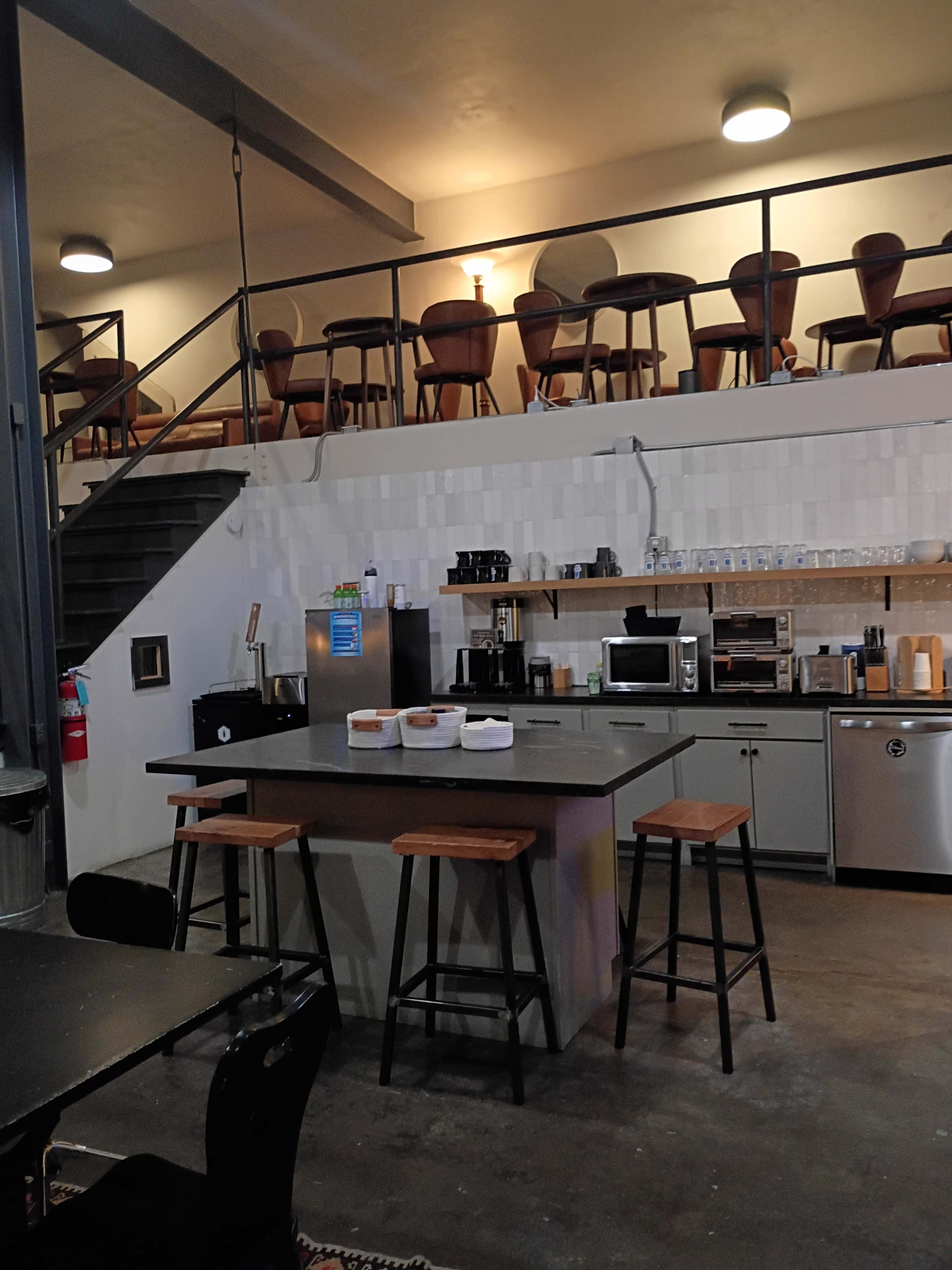 The image shows a modern kitchen area with an island, bar stools, and a staircase leading to an upper seating area with chairs.
