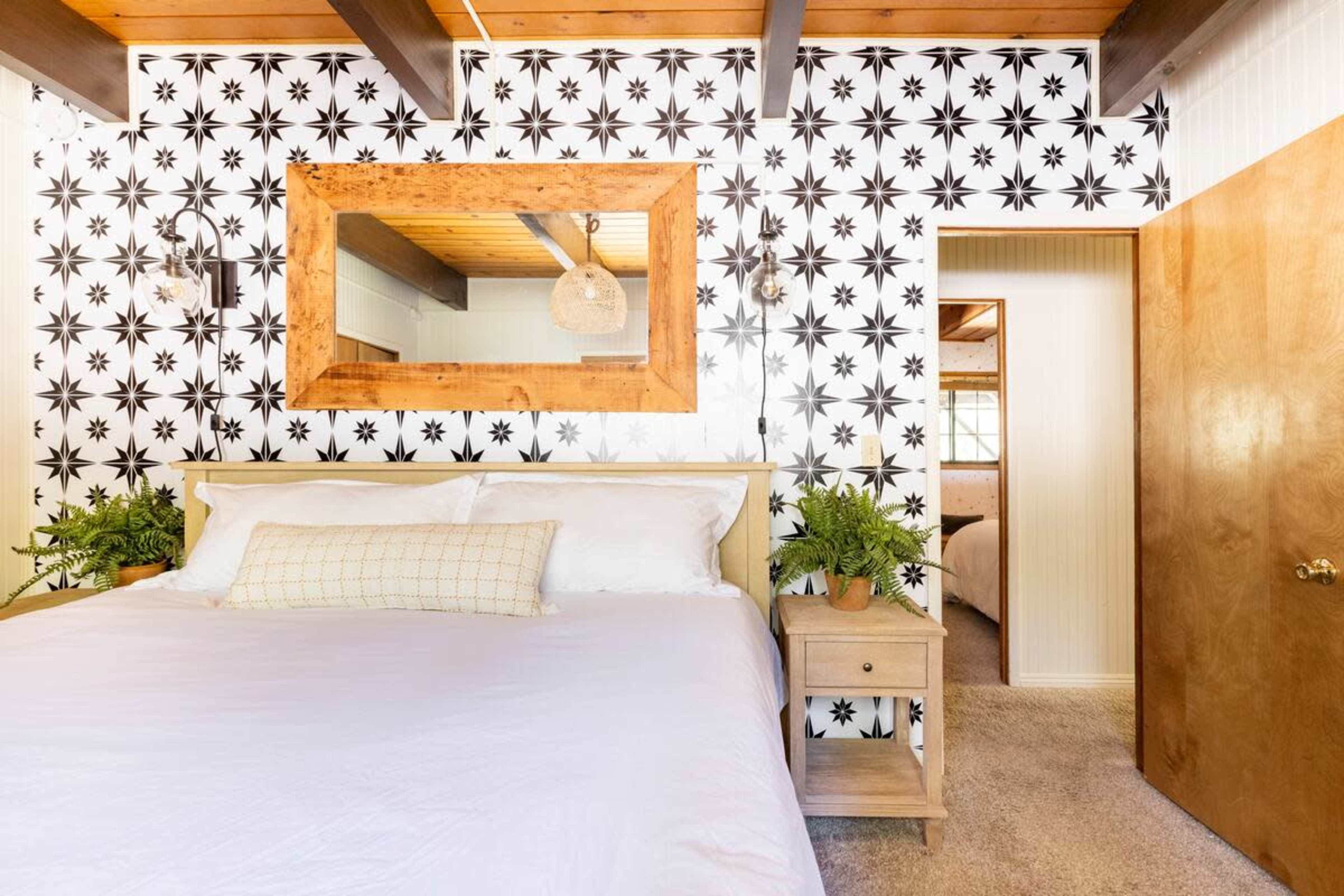 The bedroom features a large bed with white linens against a patterned black and white wall, complemented by a wooden mirror and plants on either side of a nightstand.
