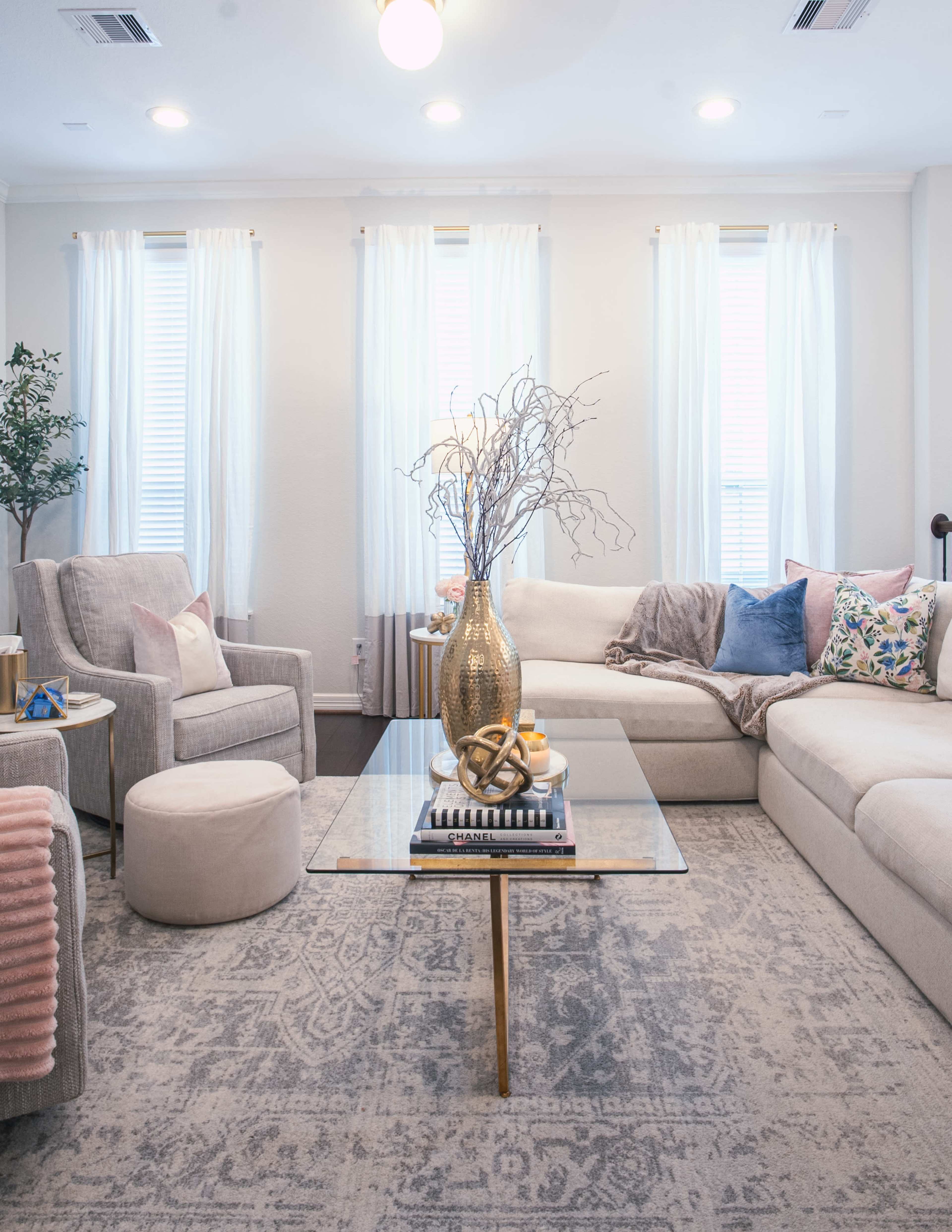 The living room features a light-colored sectional sofa, a glass coffee table with decorative items, and bright natural light streaming in from the windows.