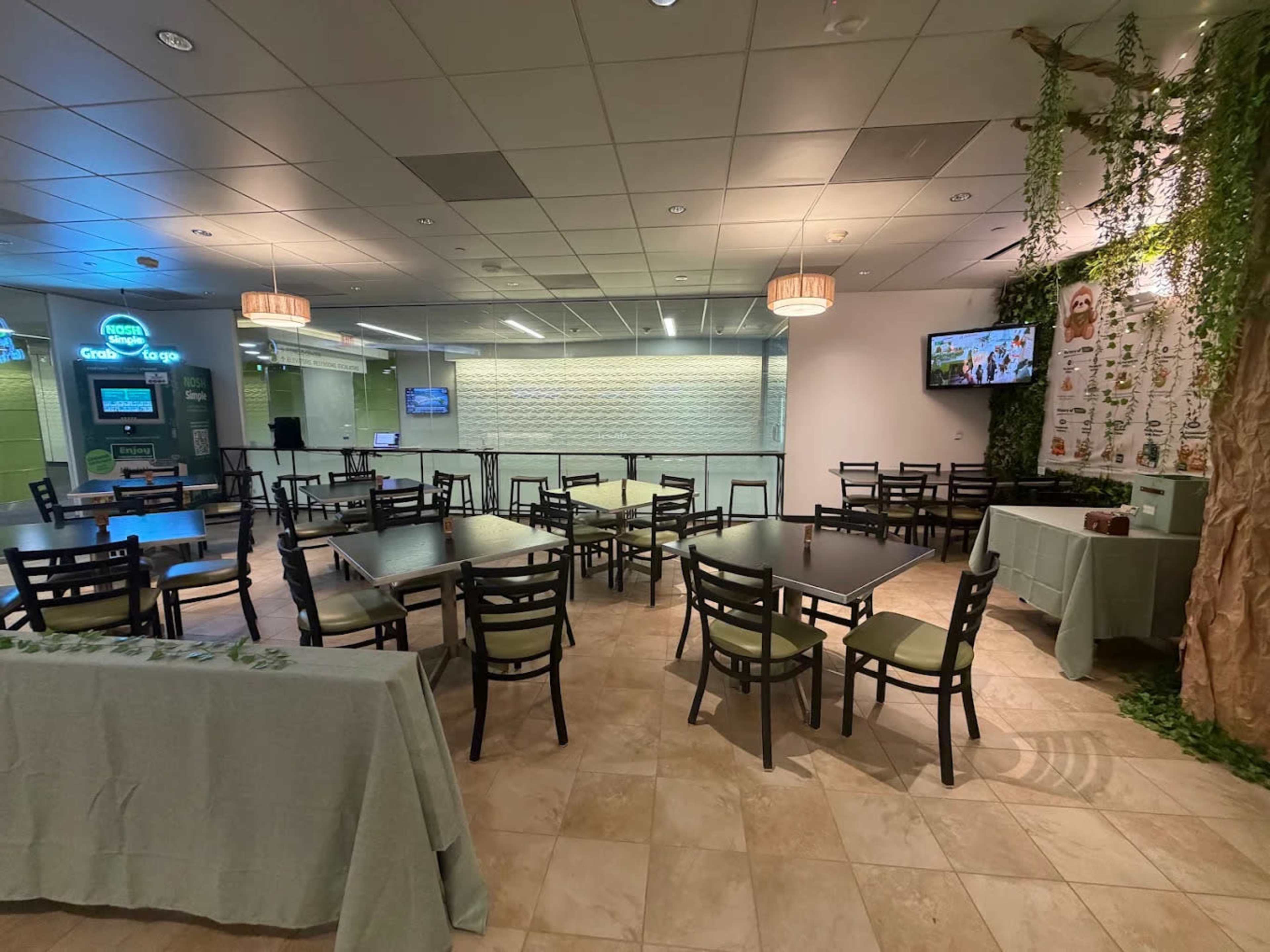 The image shows an empty dining area with several tables and chairs, featuring a green wall and a television in the background.
