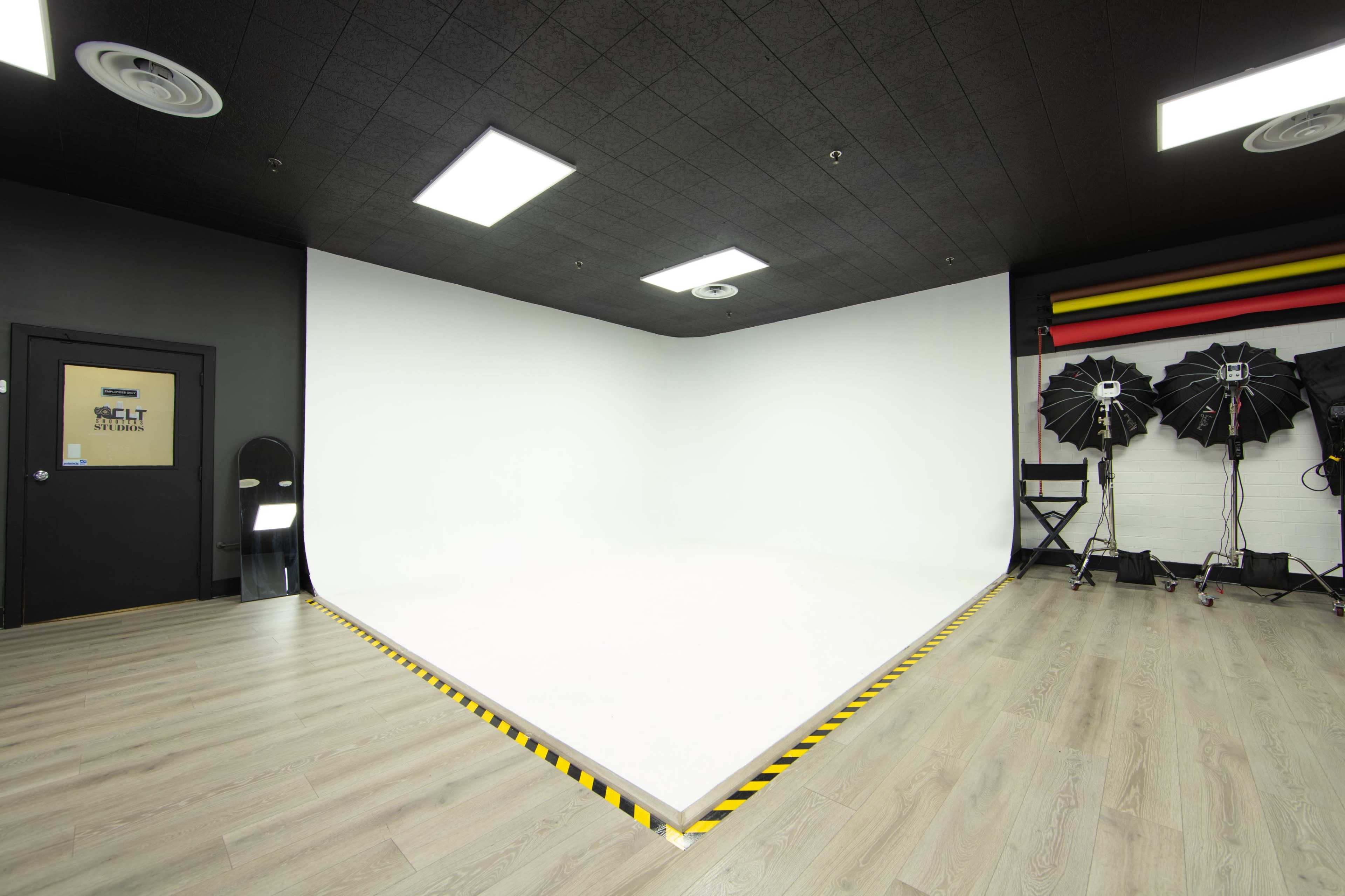 The image shows a well-lit photography studio with a seamless white backdrop, wooden flooring, and studio lighting equipment arranged in the corner.