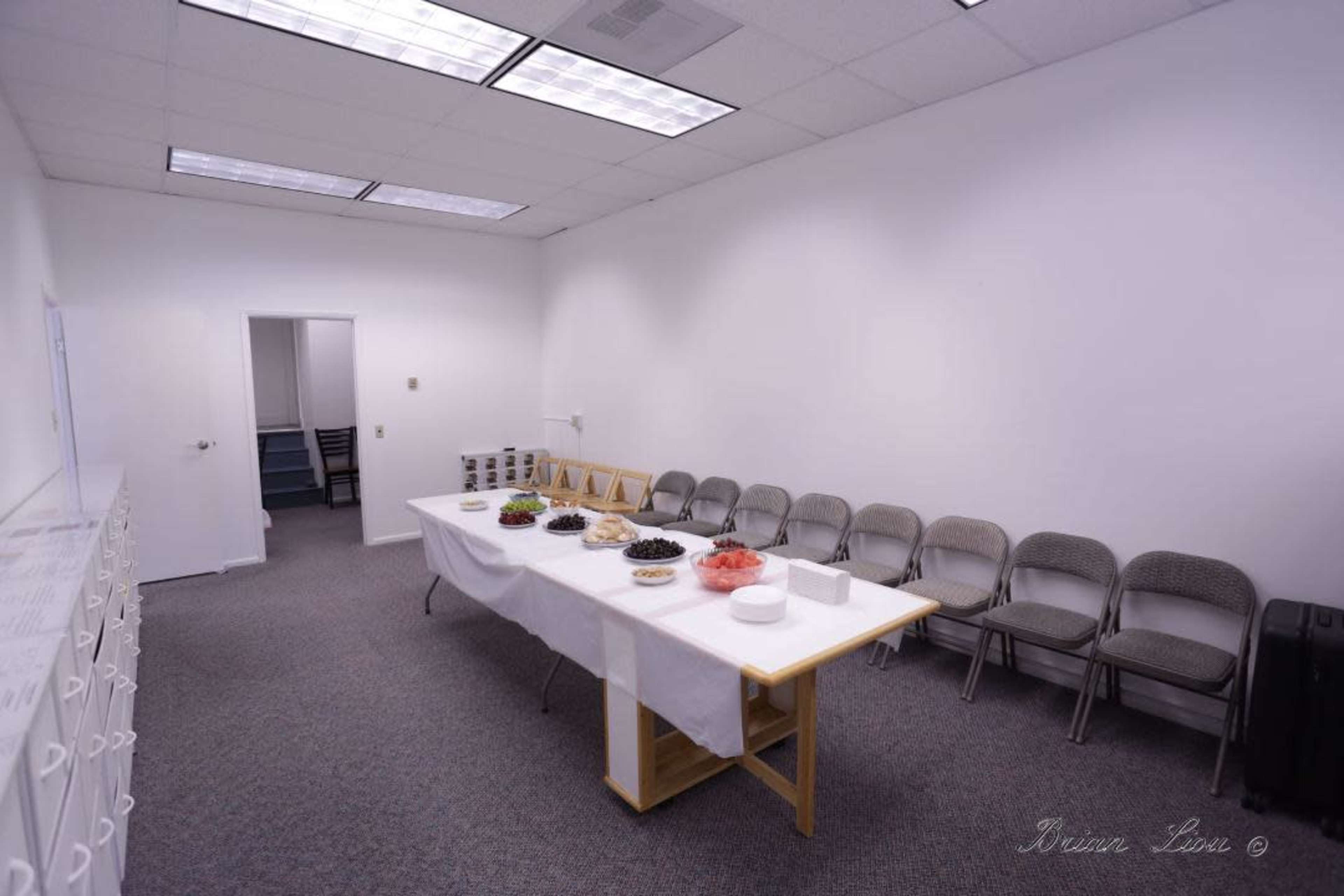 A conference room features a long table set with various foods and several gray chairs lined against the wall.