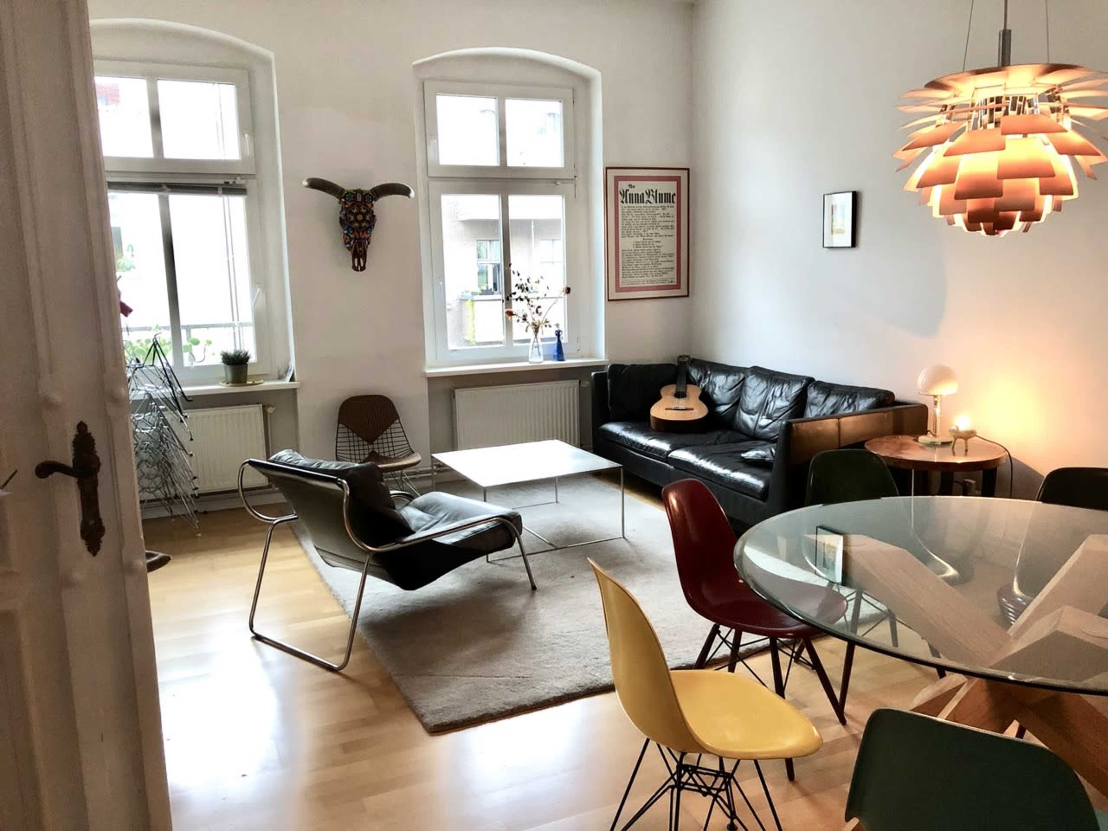 The image shows a modern living area featuring a black leather sofa, a glass dining table with colorful chairs, and an acoustic guitar on a coffee table.
