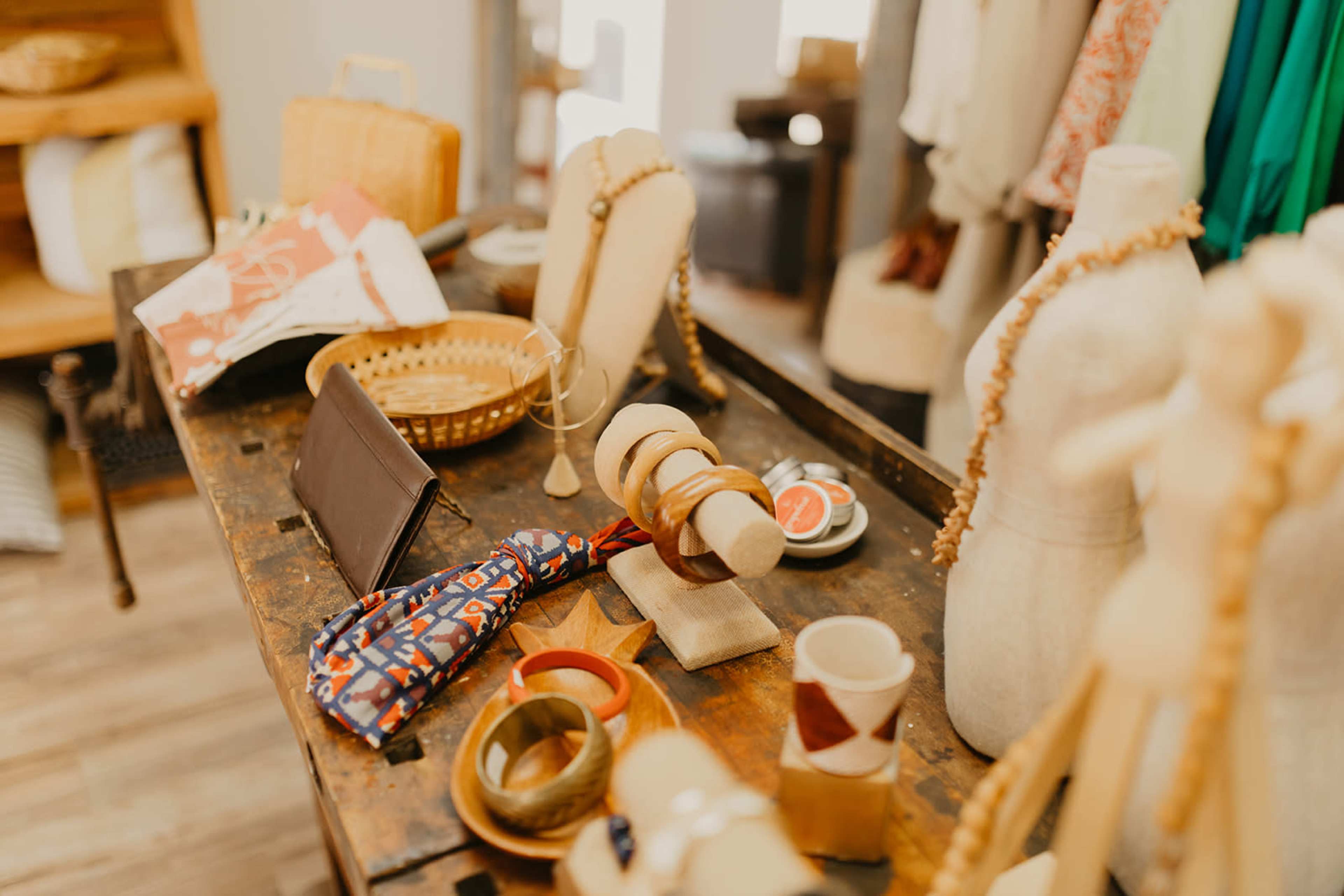A wooden table displays various accessories, including jewelry, a wallet, and a patterned cloth, in a small boutique setting.