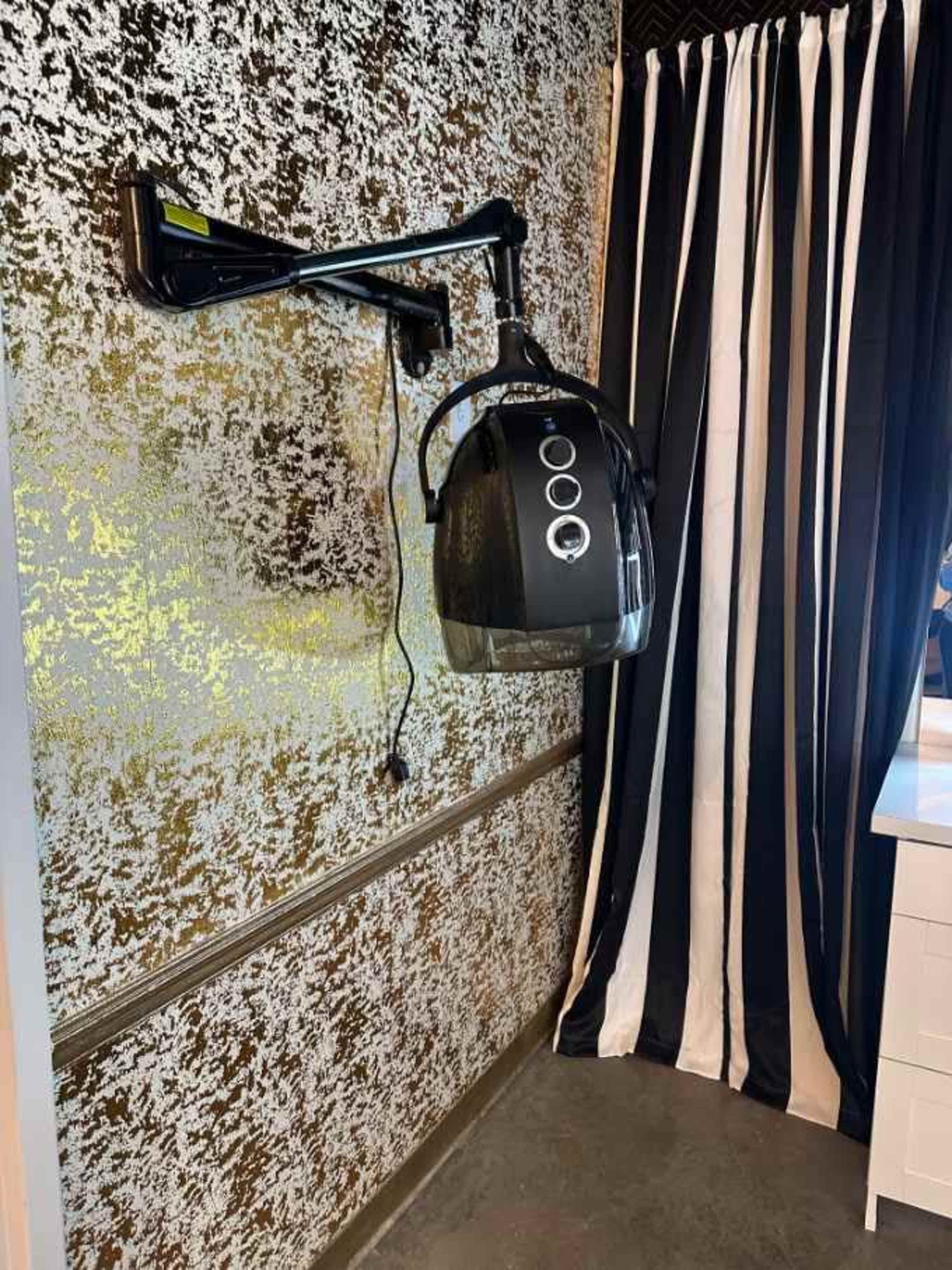 A wall-mounted hairdryer hood is attached to a patterned gold and silver wall, next to a black and white striped curtain.