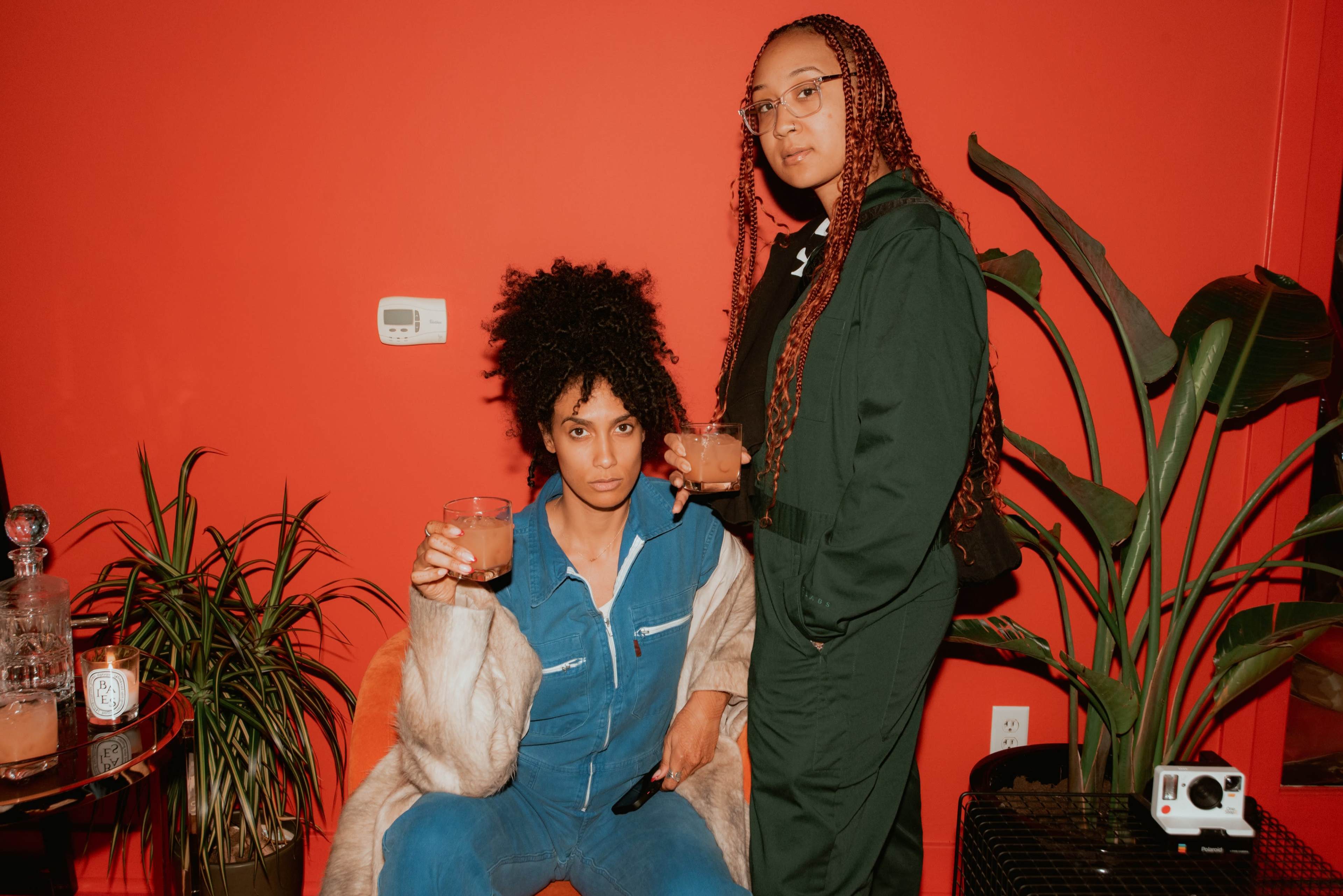 Two women hold drinks while posing in front of a vibrant red wall, surrounded by indoor plants and furniture.