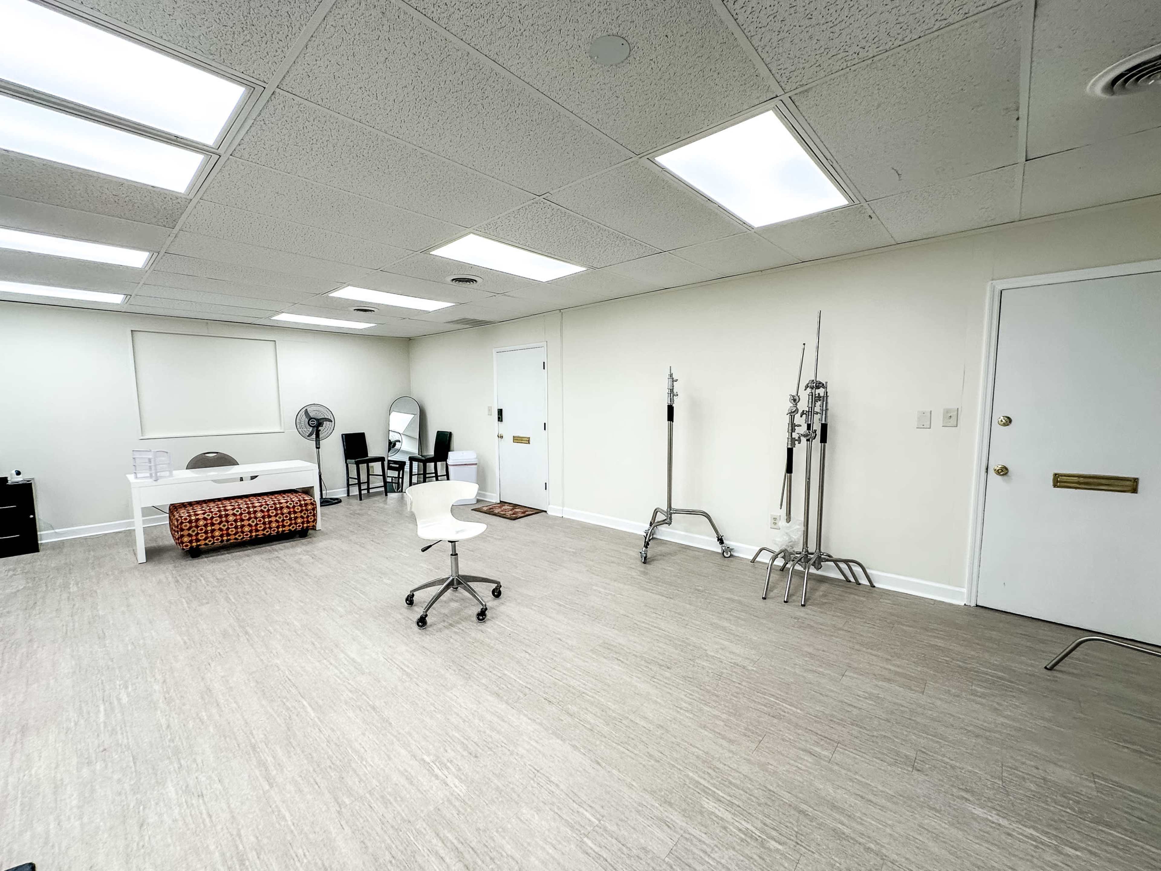 A minimalistic, empty room with beige walls, fluorescent lighting, a white chair on wheels, a vanity mirror, and several medical stands along one wall.