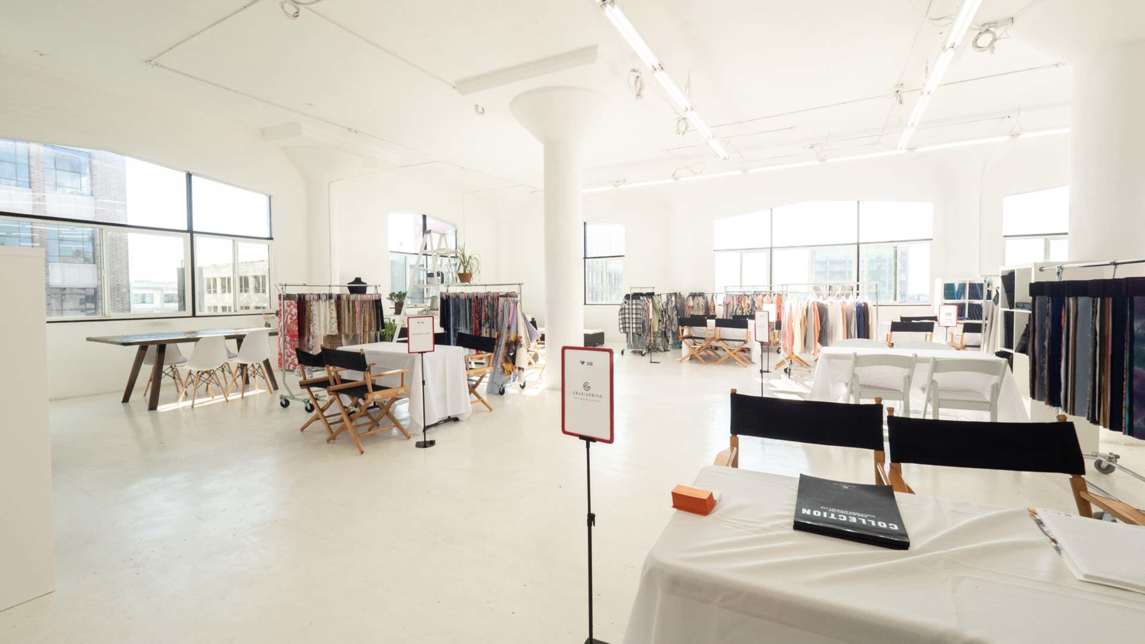 A bright, spacious room features multiple display racks filled with various fabric samples and folding chairs arranged around tables.