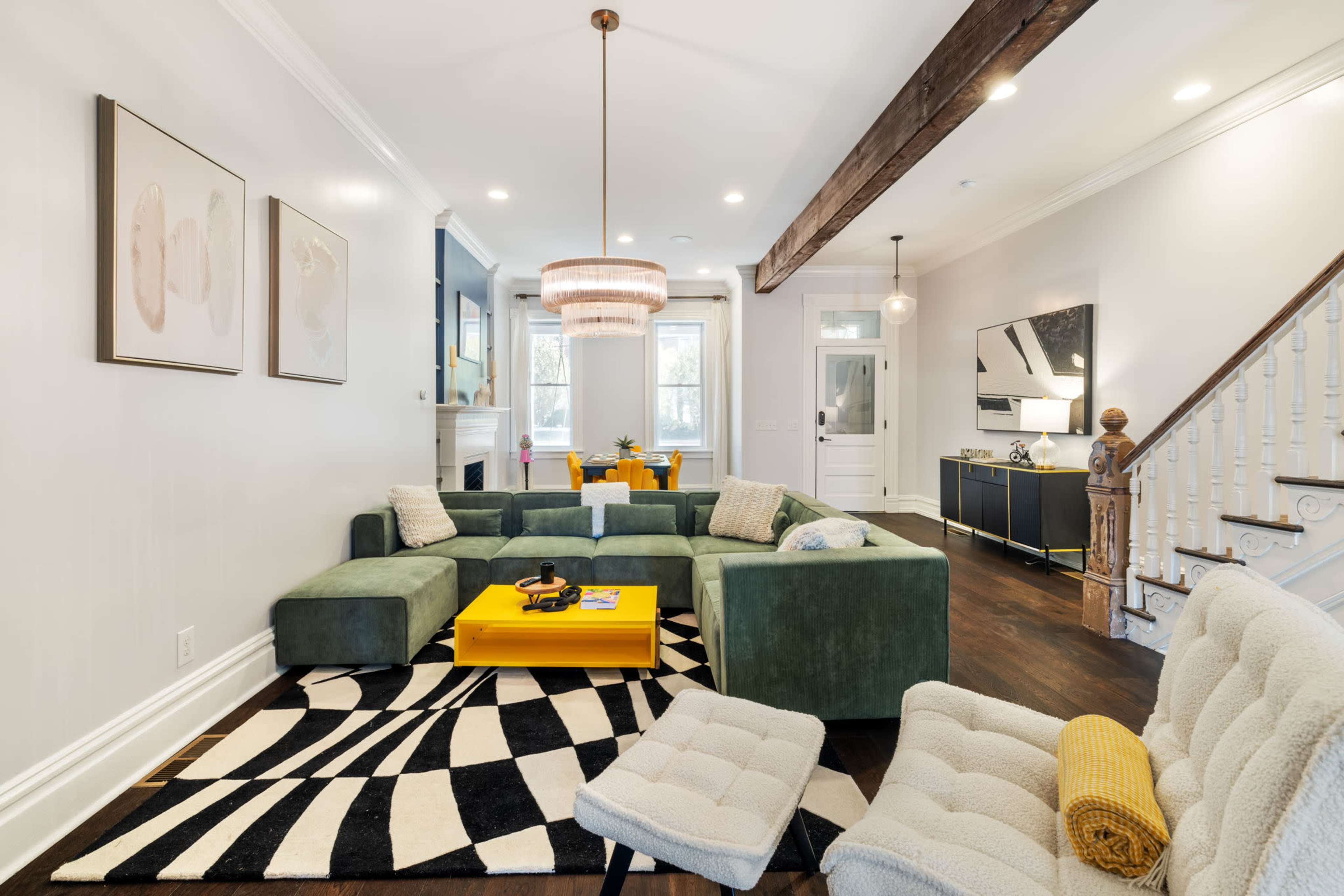 A modern living room featuring a green sectional couch, a yellow coffee table, and a patterned black and white rug, with a light fixture and decorative art on the walls.