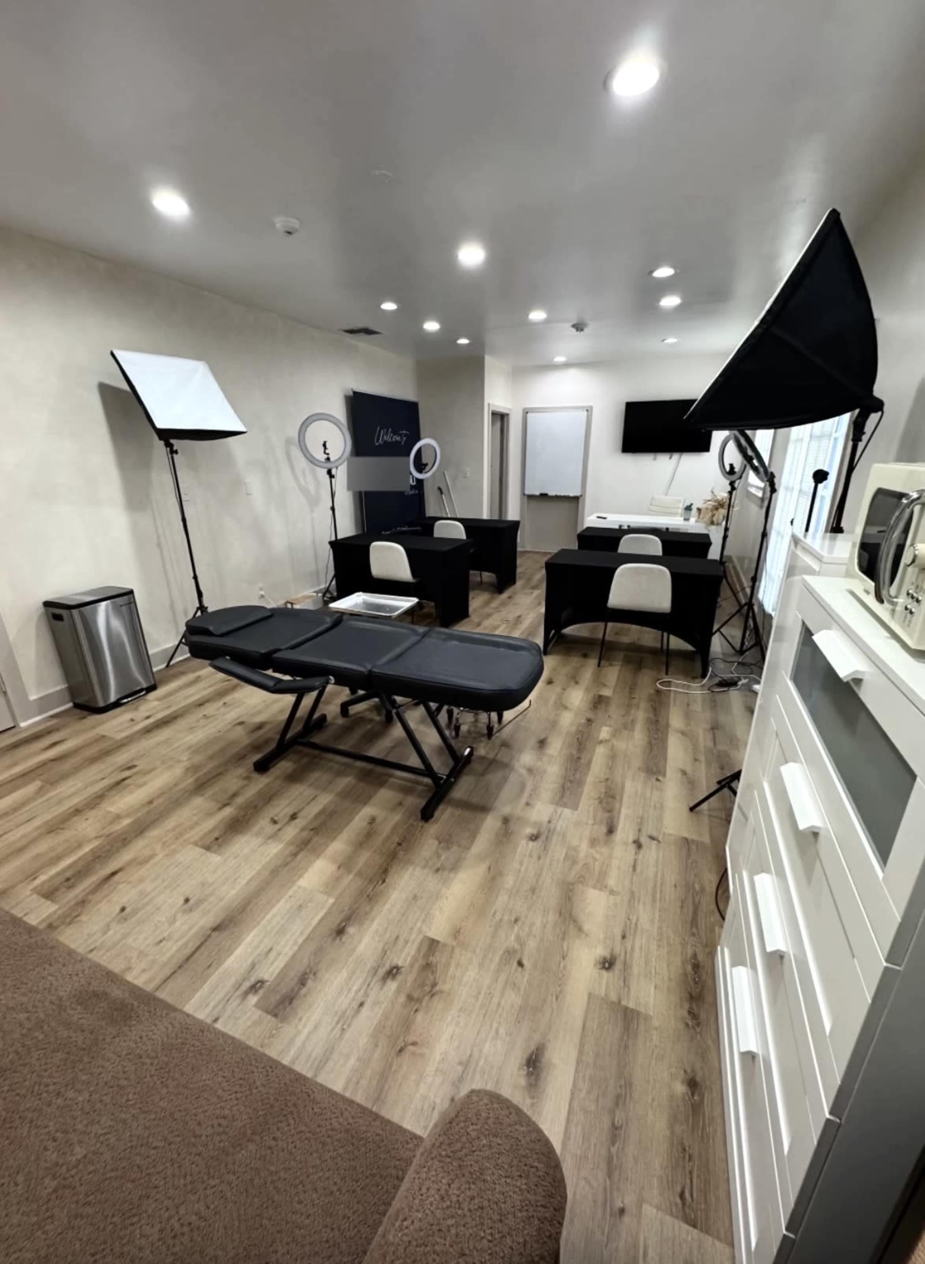 The image shows a well-lit room with wooden floors, featuring two treatment tables, a desk, several photography lights, and a white storage cabinet.