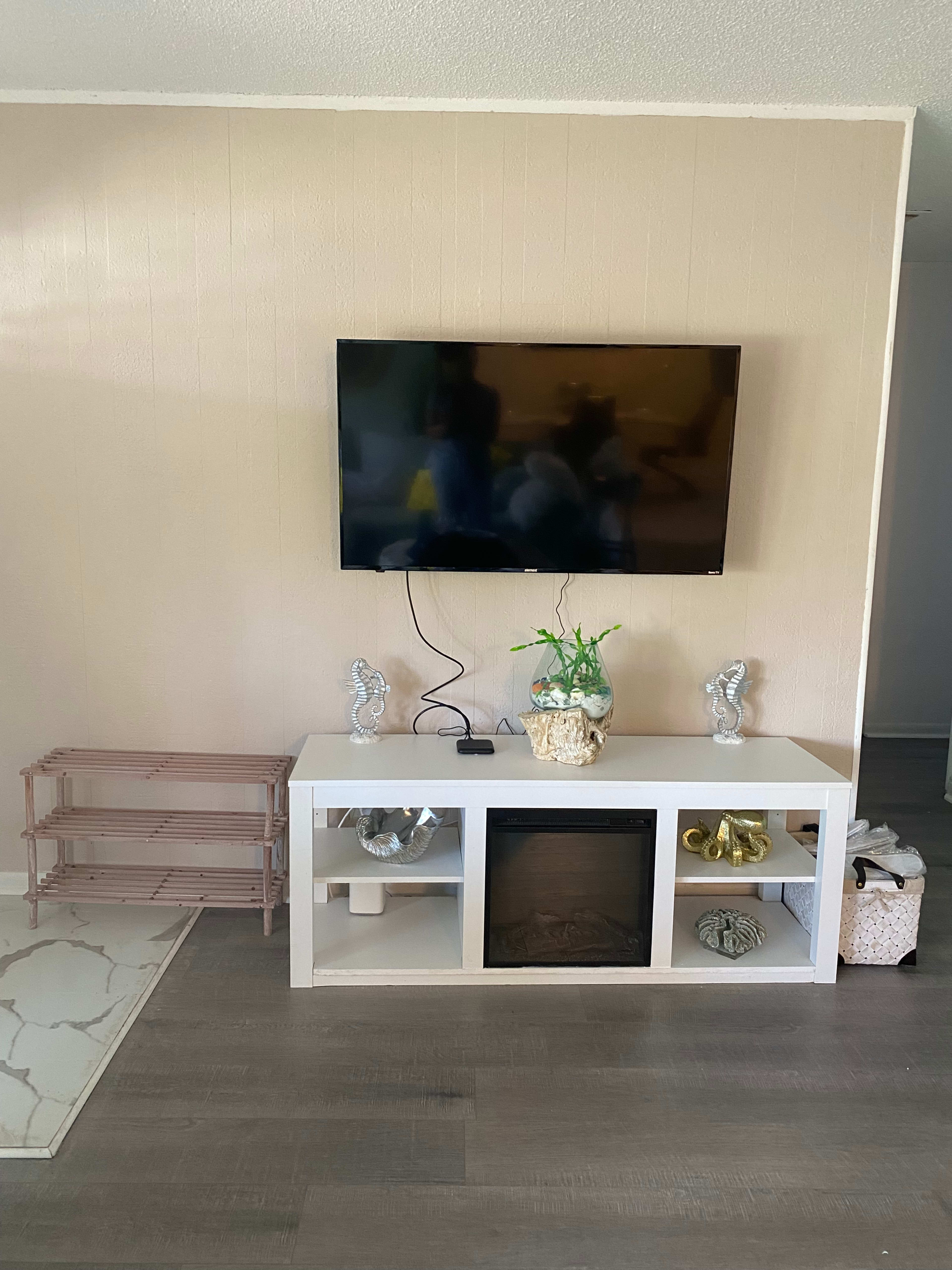 A modern living room setup featuring a wall-mounted television above a white entertainment unit with decorative items and a small refrigerator.