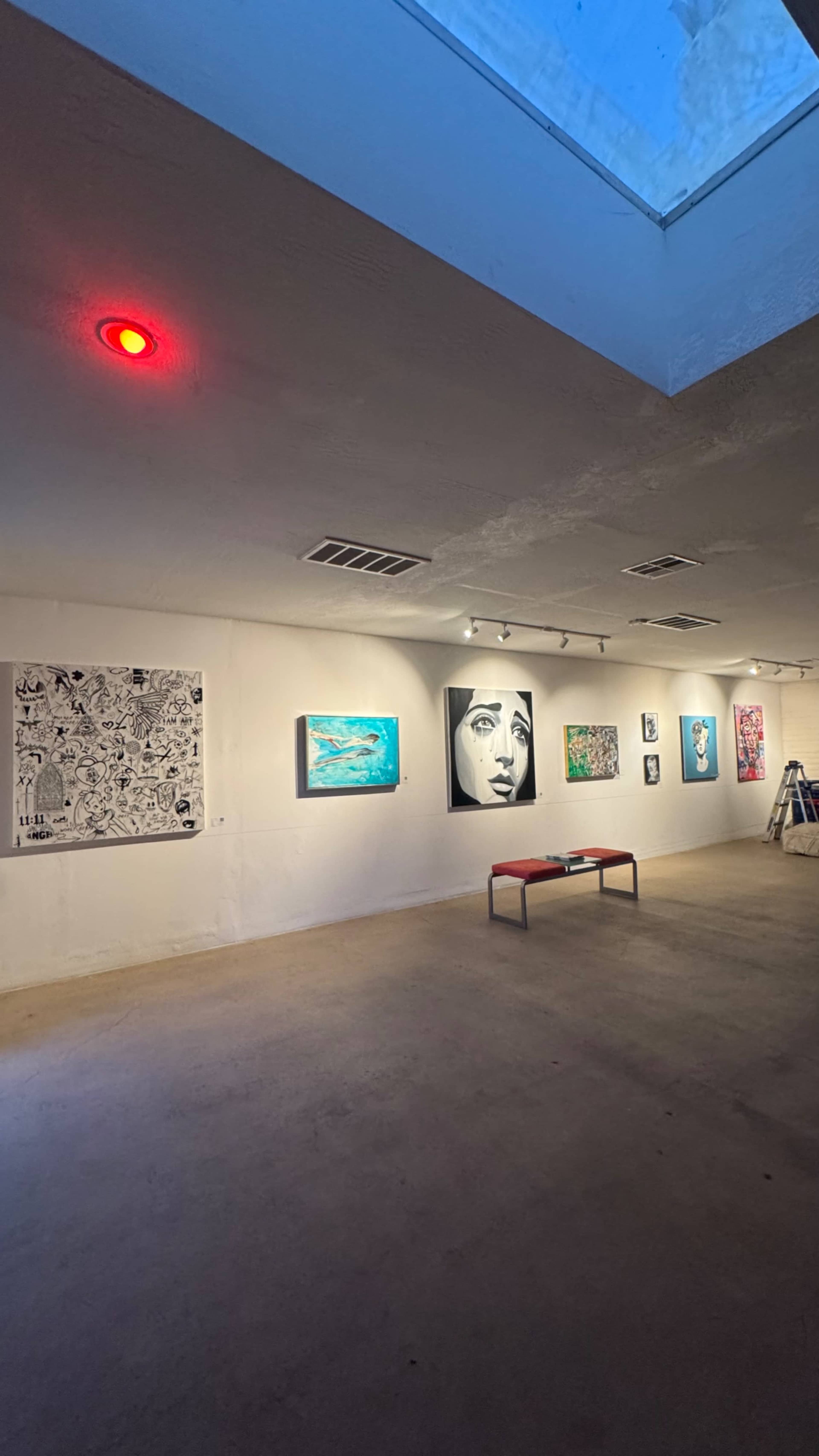 The image shows an art gallery featuring several paintings on the walls, with a bench positioned in the center and a skylight above.