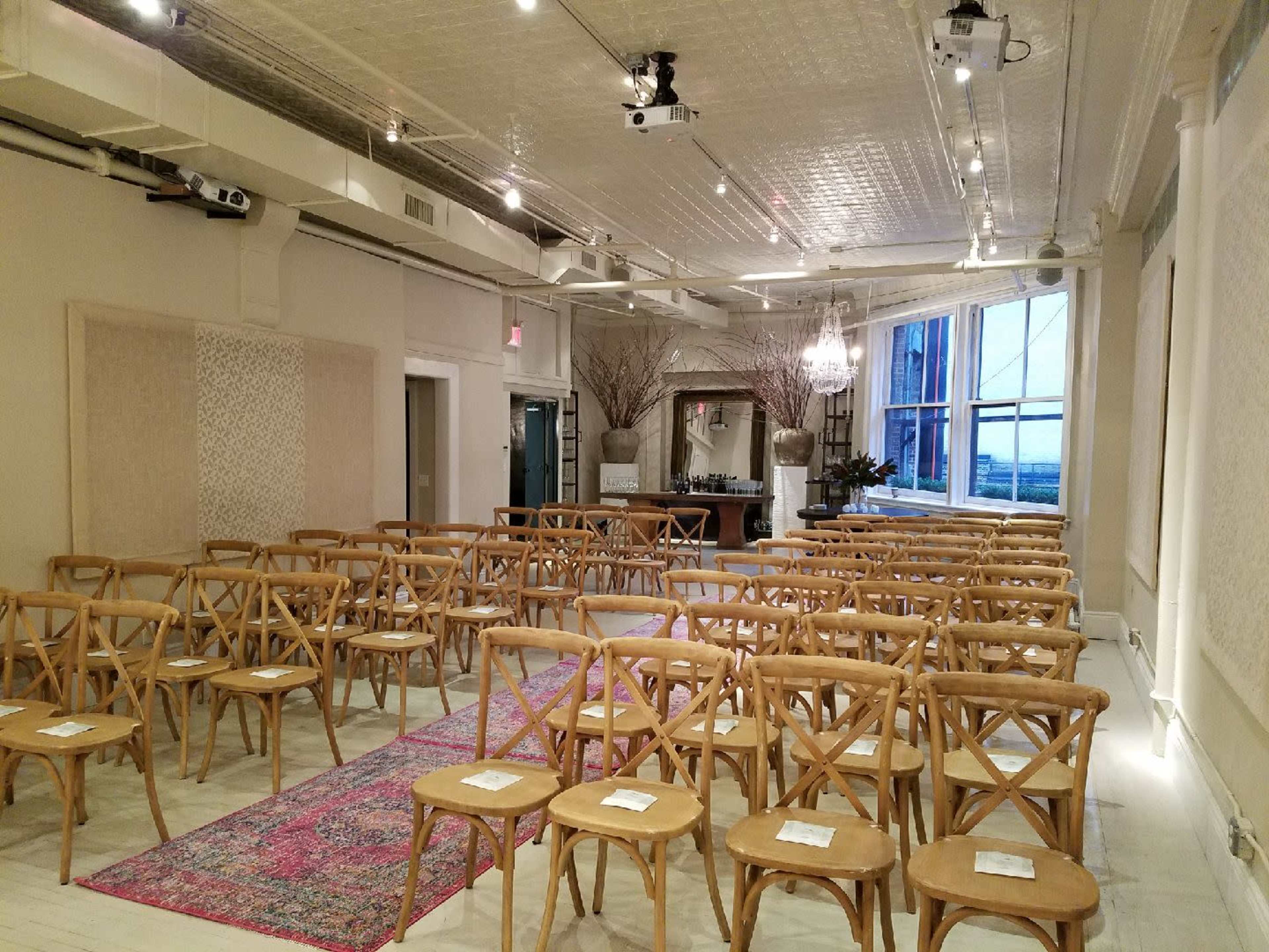 A spacious room is set up for an event, featuring rows of wooden chairs arranged on a decorative rug, with large windows offering a view of the outside.