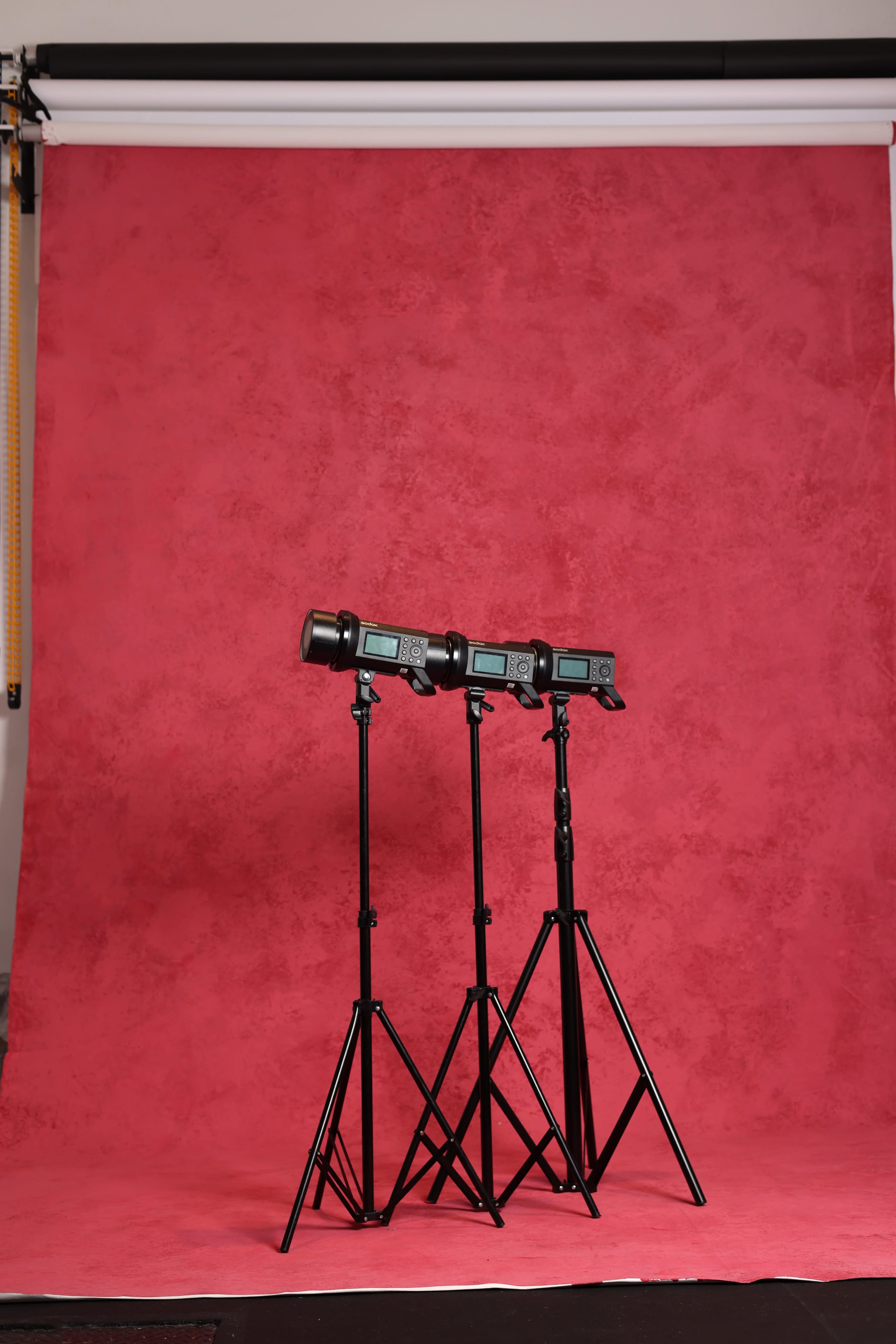 Three studio lights on black tripods are positioned in front of a textured red backdrop.