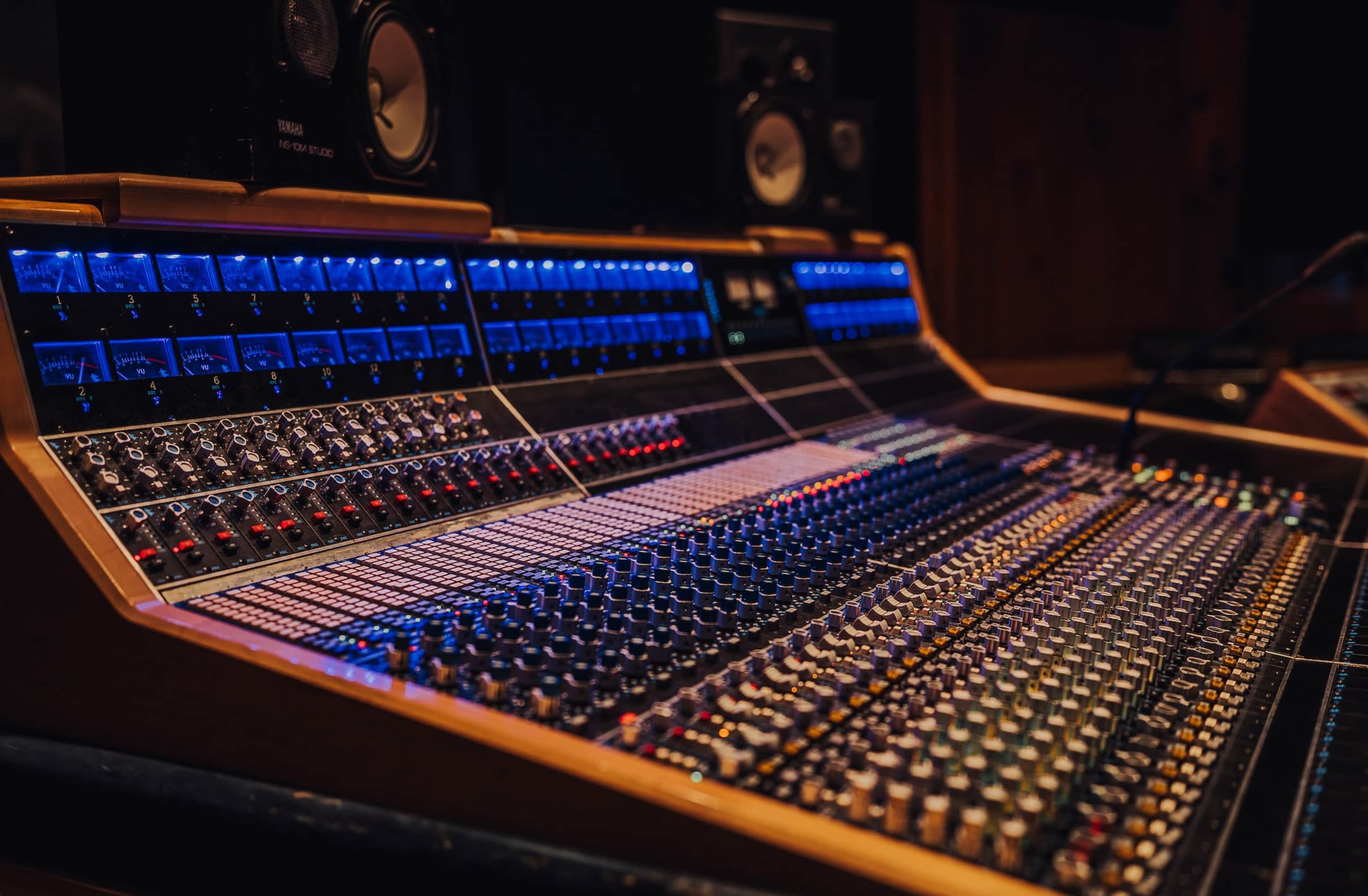 The image shows a large audio mixing console with numerous colorful controls, knobs, and buttons, set in a dimly lit studio environment.