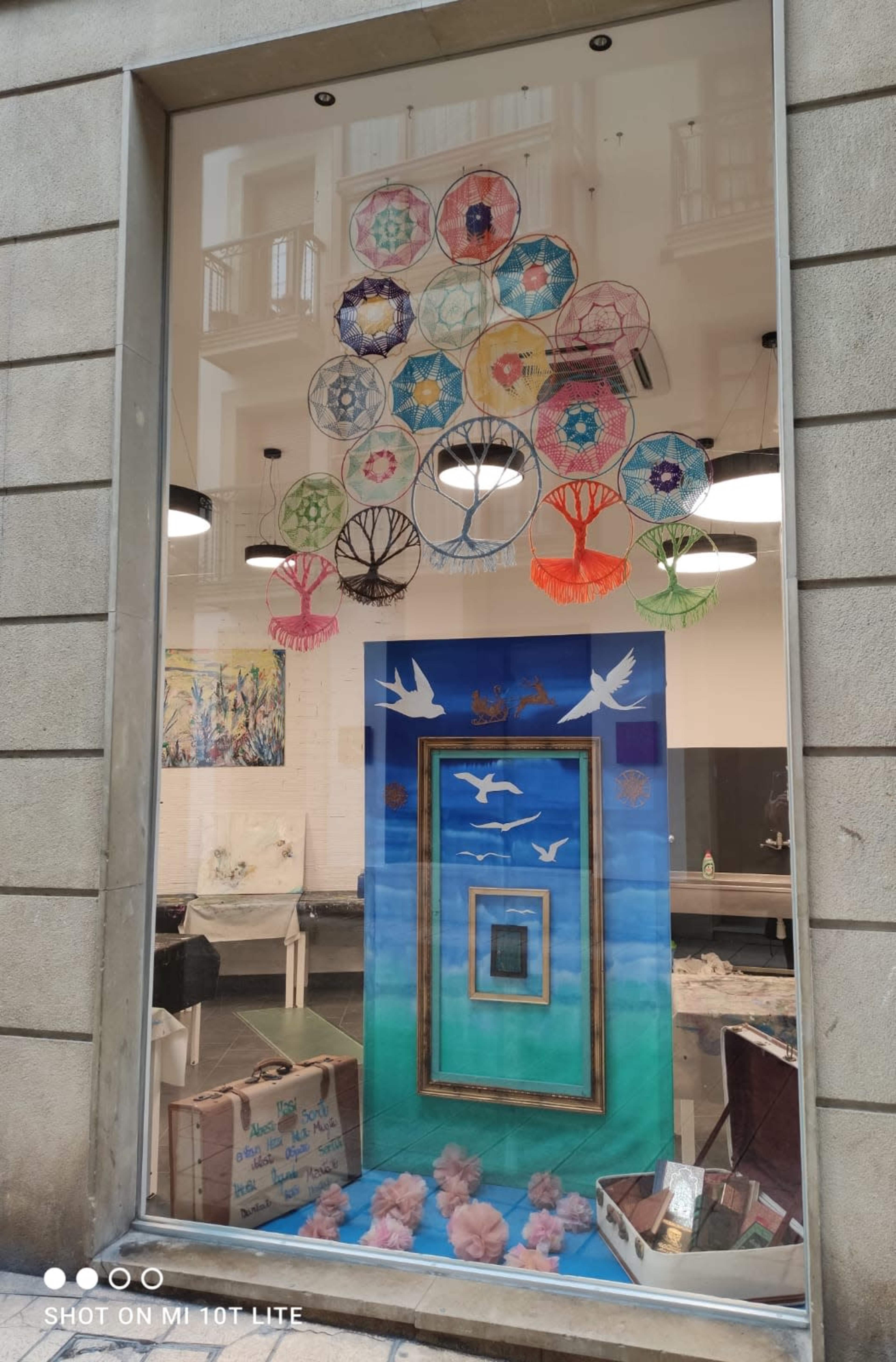 The image shows a shop window display featuring colorful decorative umbrellas hanging above a blue backdrop with white birds and a framed picture.