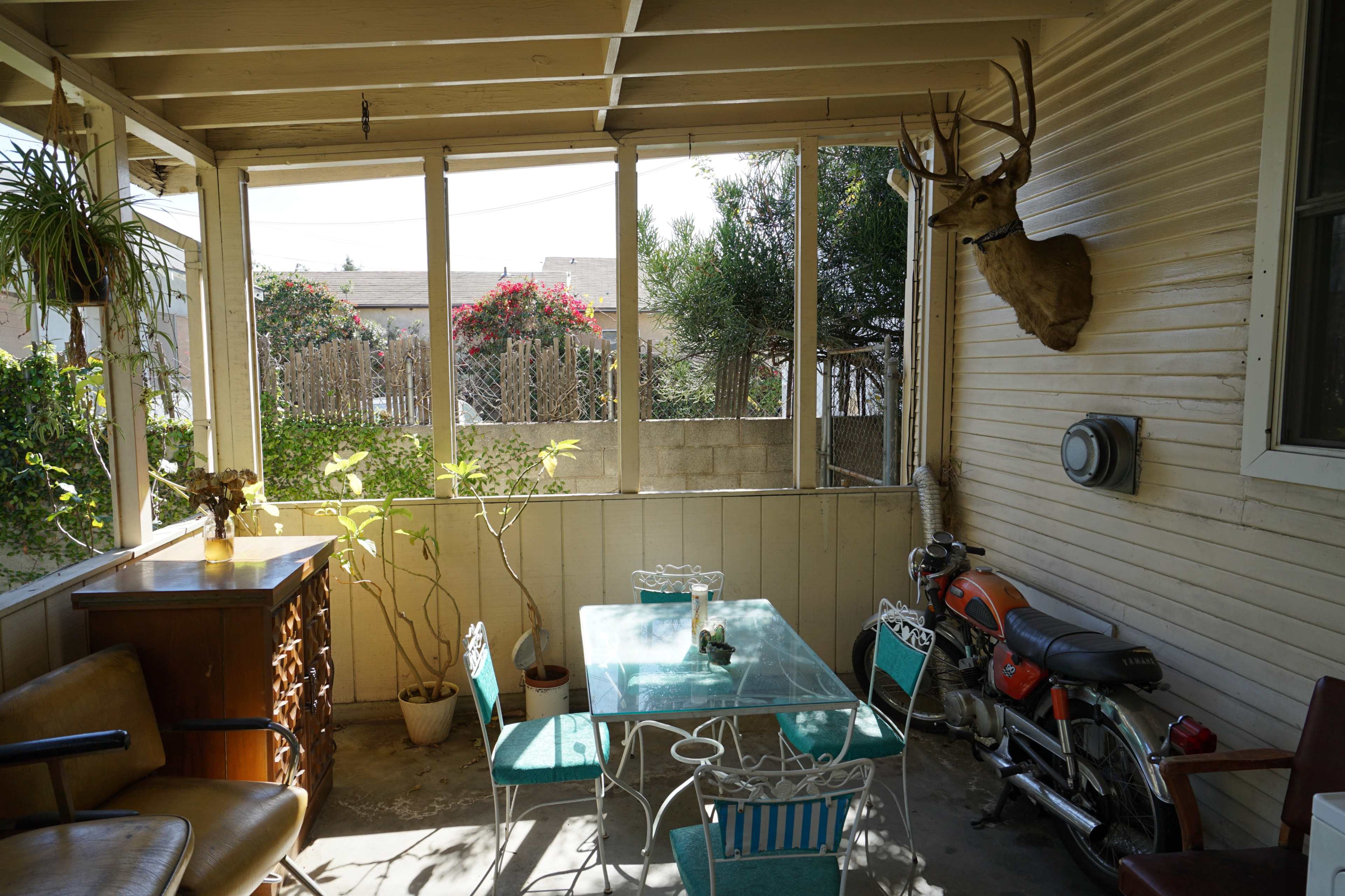 A screened porch with a glass table and chairs, a wooden cabinet, a mounted deer head on the wall, and a motorcycle parked nearby, surrounded by plants and a garden view.