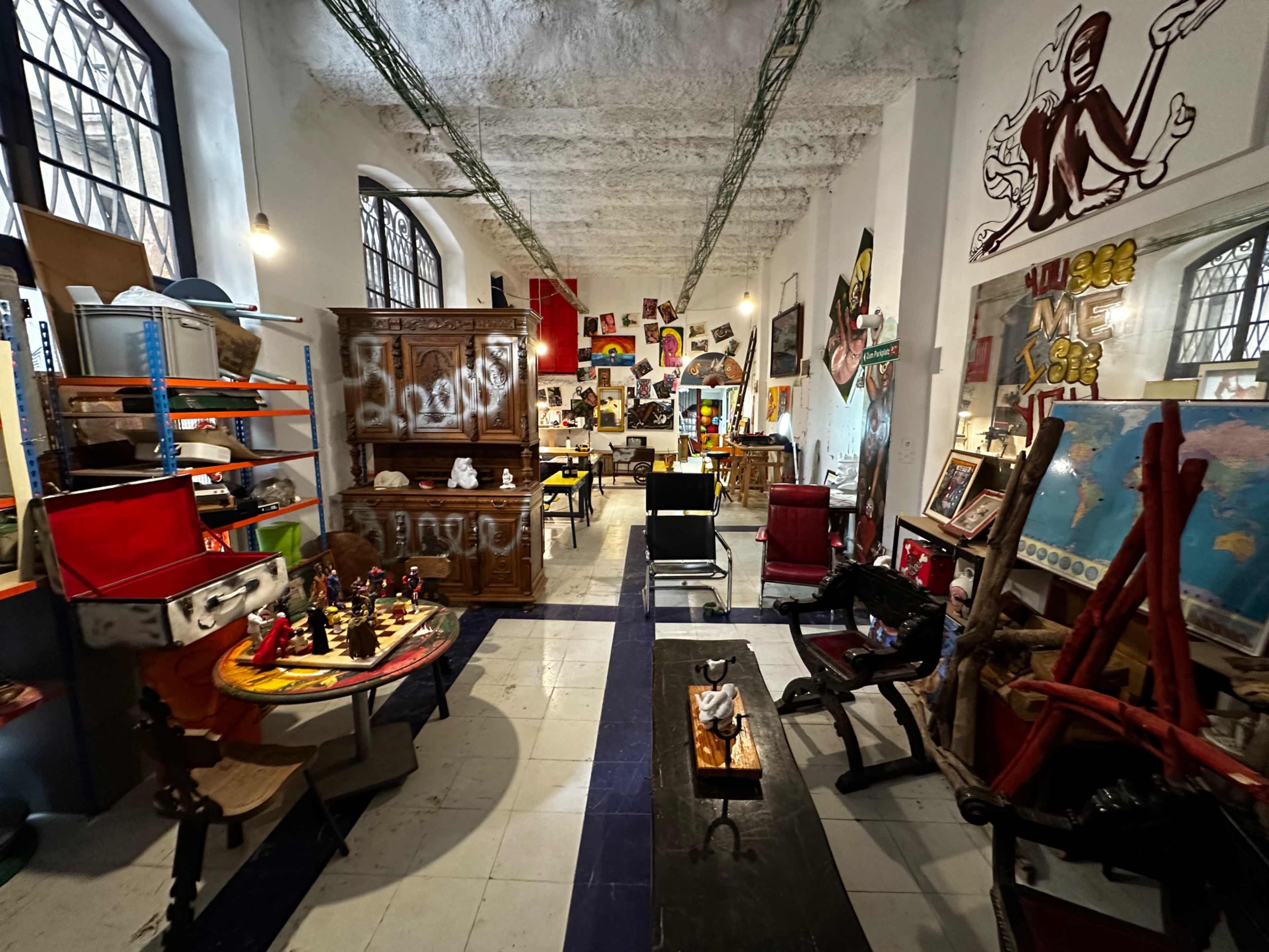 The image shows a vibrant art studio filled with various pieces of furniture, artwork, and sculptures, with colorful murals on the walls.