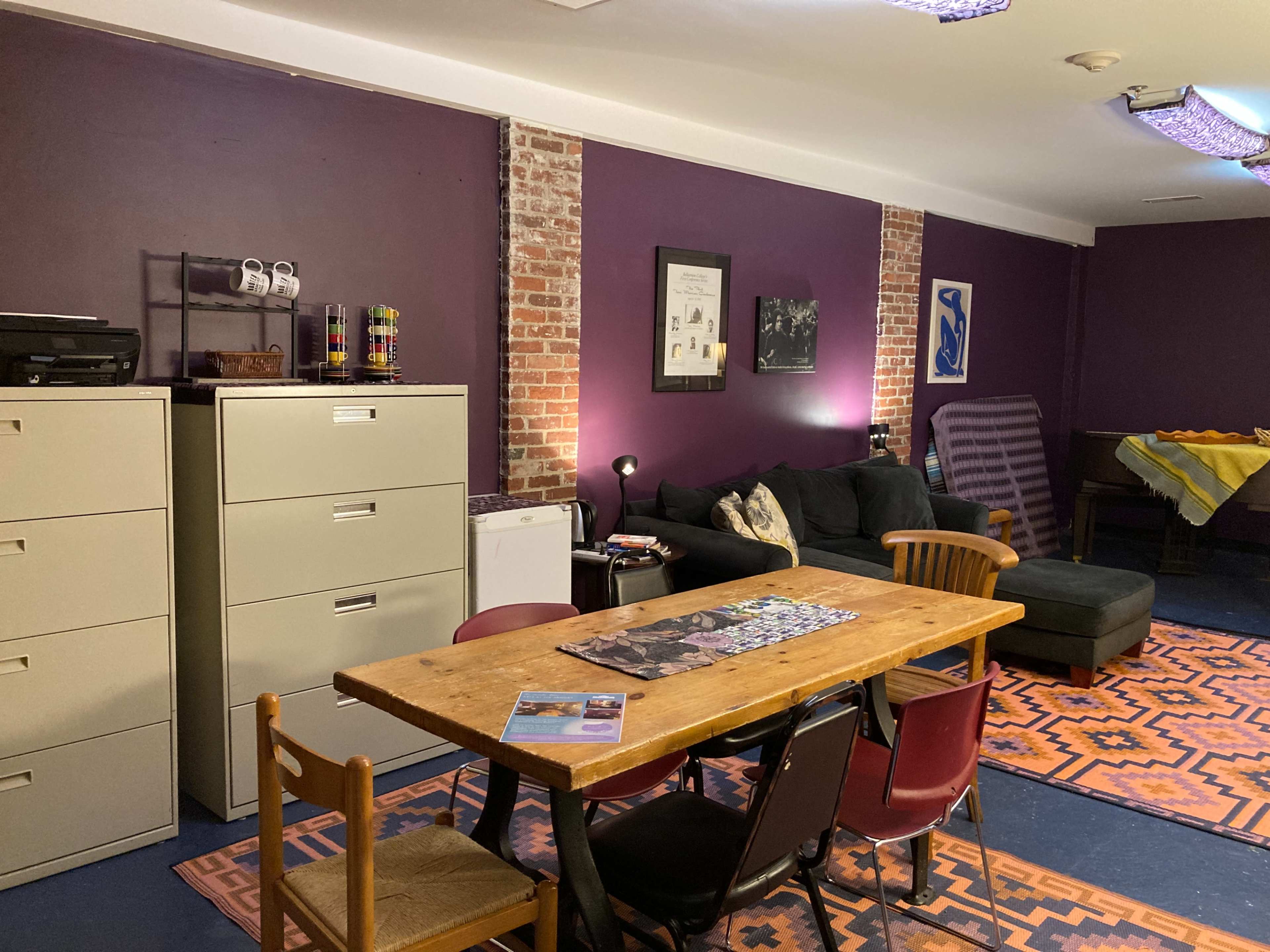 A room with purple walls, a wooden table surrounded by chairs, filing cabinets, a couch, and a patterned rug.