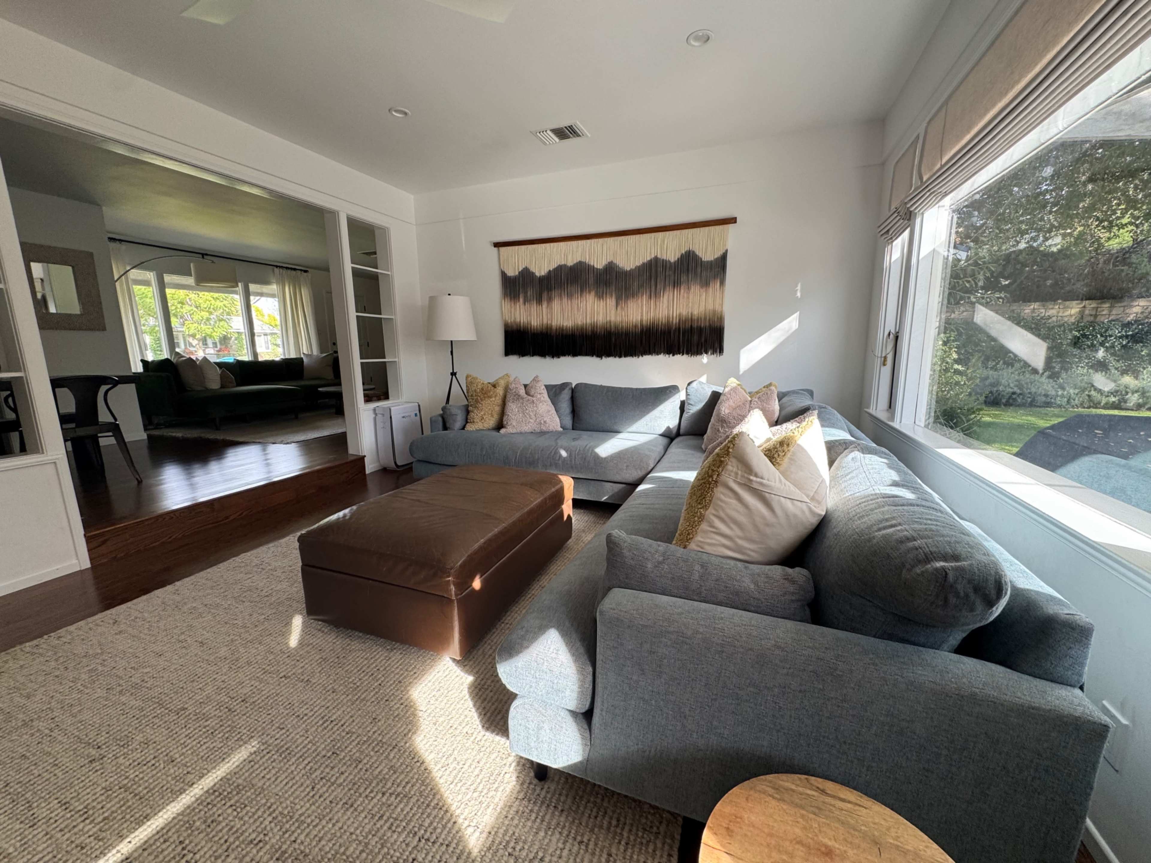 The image shows a bright living room with two couches, a coffee table, and a textured wall hanging, overlooking a garden area through large windows.