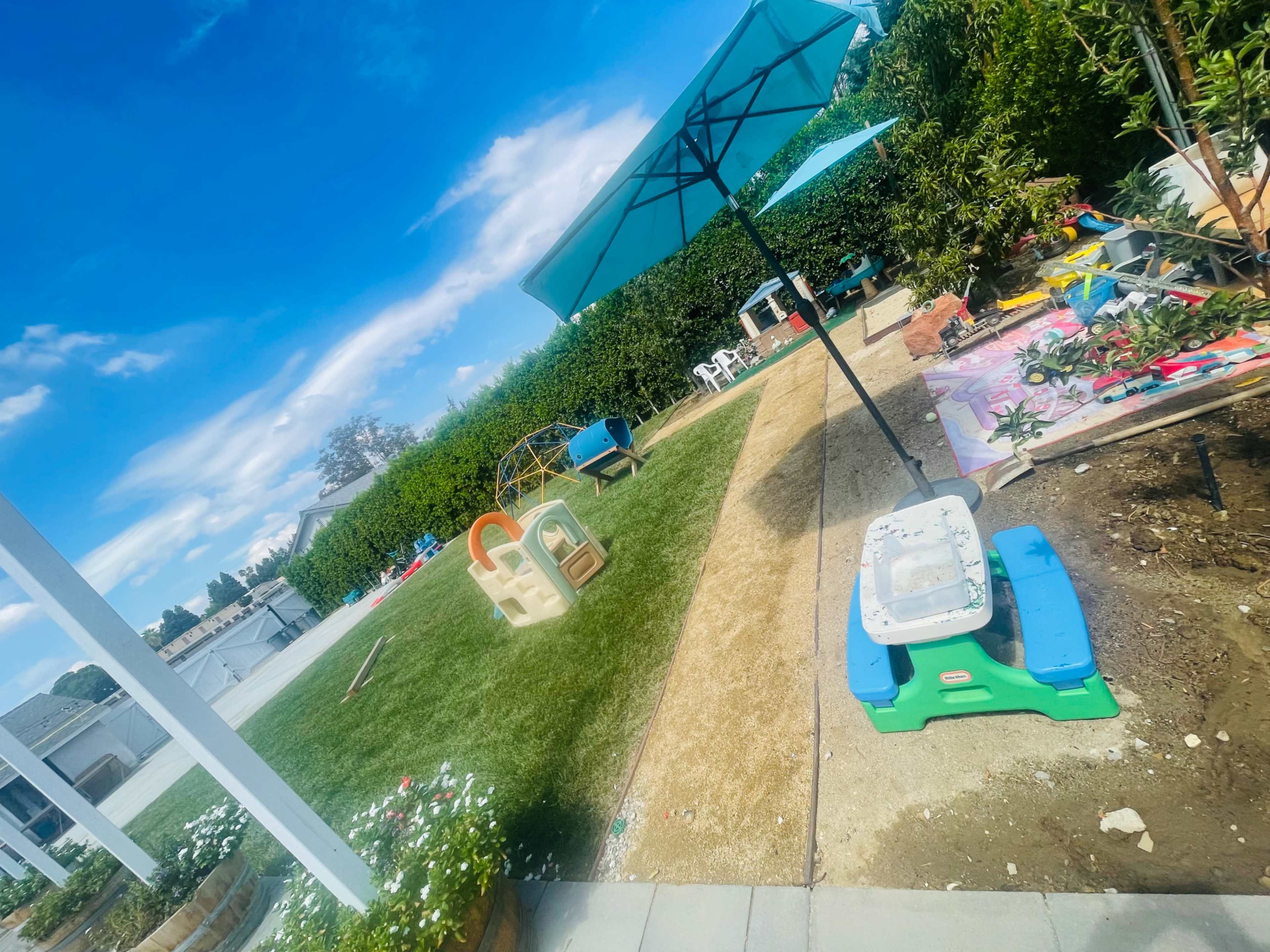 A backyard scene features a green lawn, a sandbox, a playhouse, and patio furniture under two umbrellas.