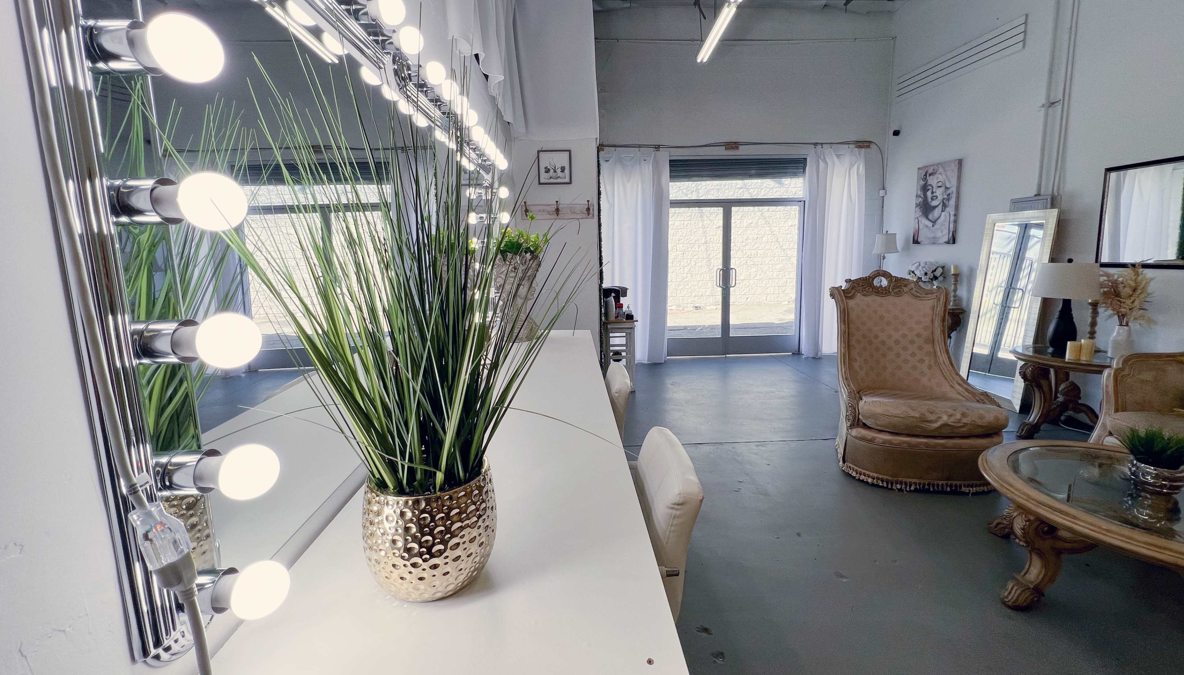 The image shows a bright, spacious vanity area with a row of lighted mirrors and a potted plant on a countertop, leading to an adjacent seating area with a decorative chair and a table.