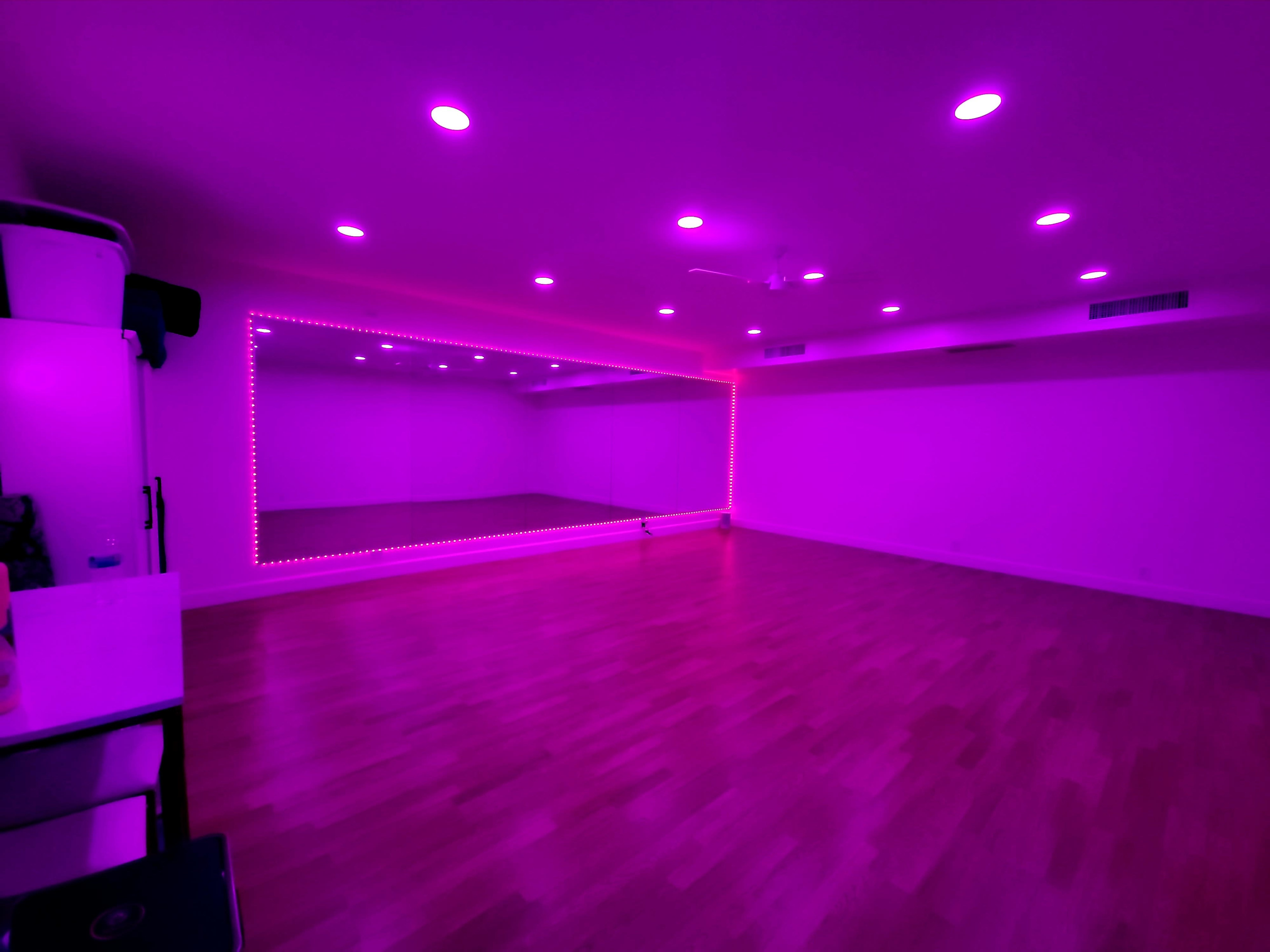 A spacious, empty room with a wooden floor, illuminated by pink LED lights and featuring a large mirror on one wall.