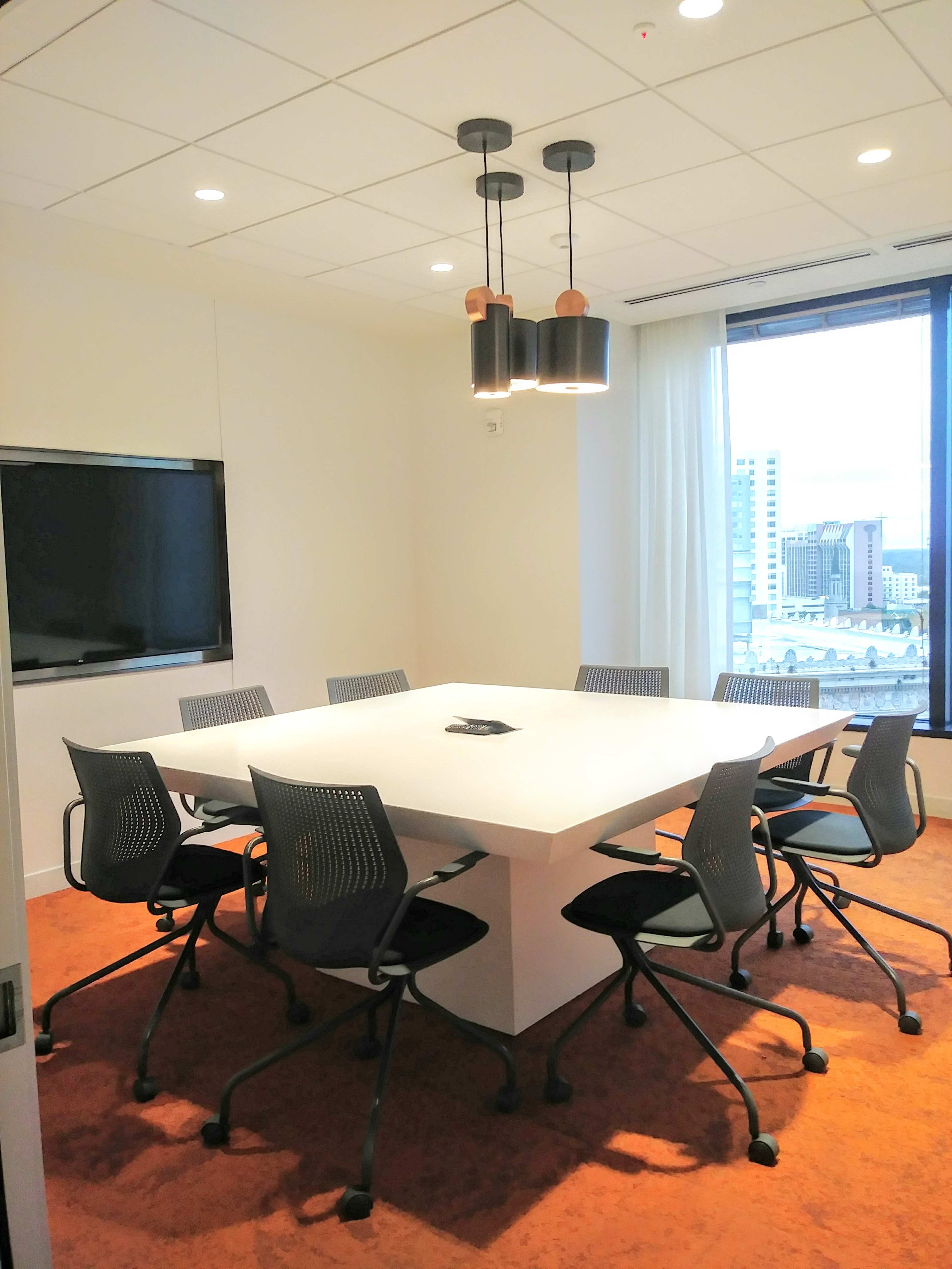 A modern conference room features a large white table surrounded by eight ergonomic chairs, with a television mounted on the wall and large windows providing a view of the city.