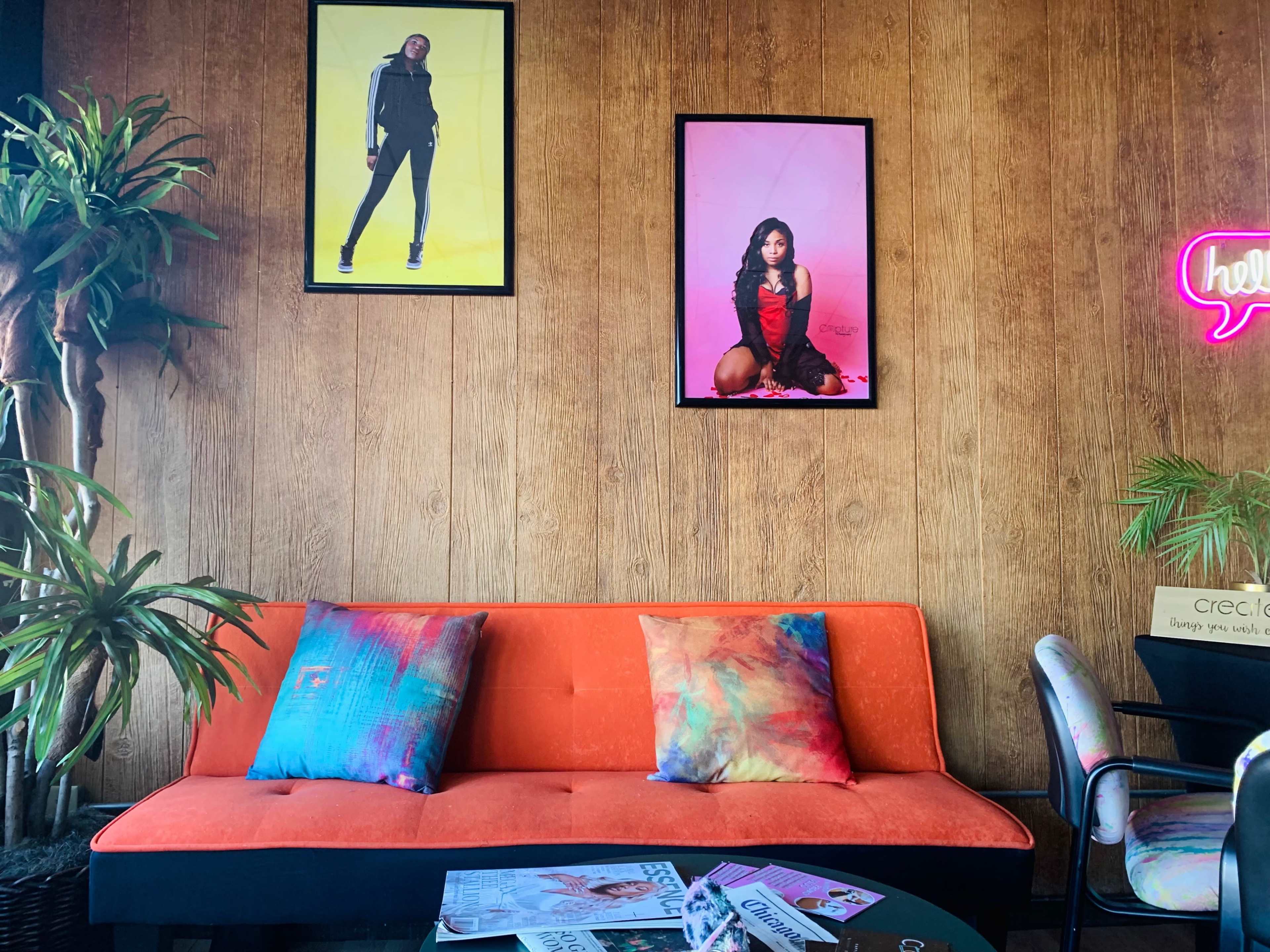 The scene features an orange sofa with colorful pillows, flanked by two framed fashion photographs on a wooden wall, alongside plants and a neon sign.