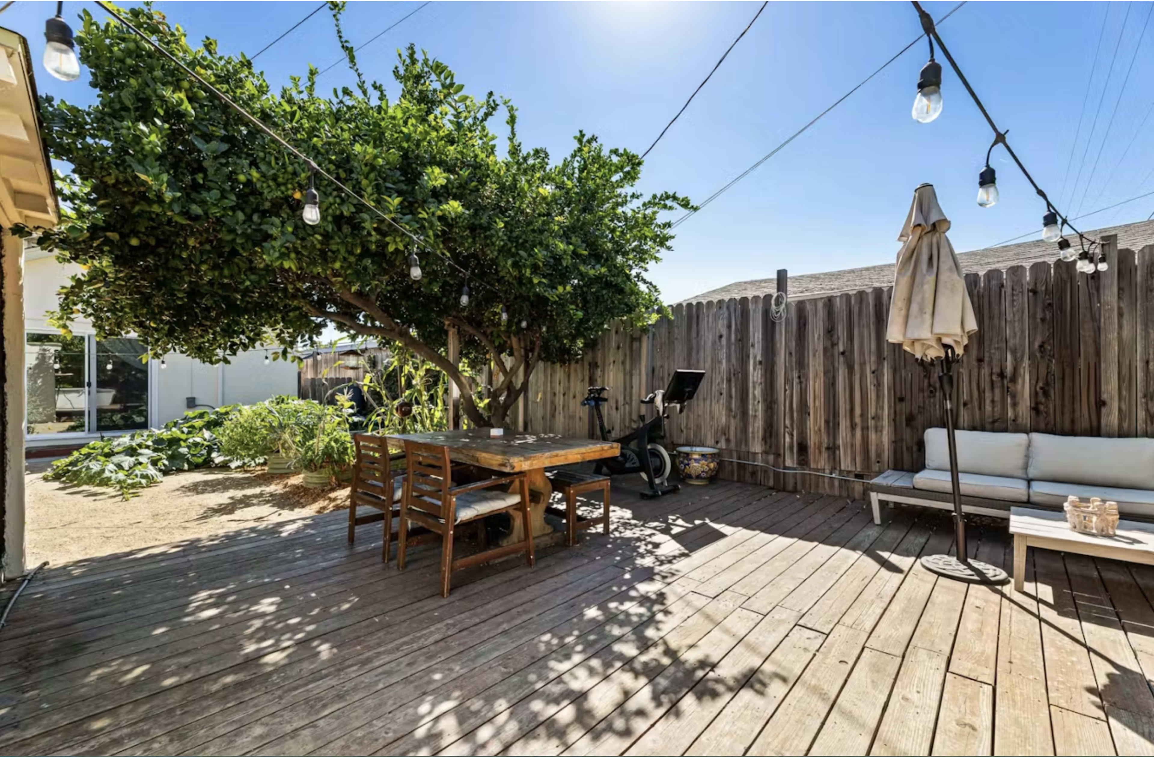 The image shows a wooden deck in a backyard with a large tree, a dining table set, a lounge chair, and hanging string lights, surrounded by a wooden fence.