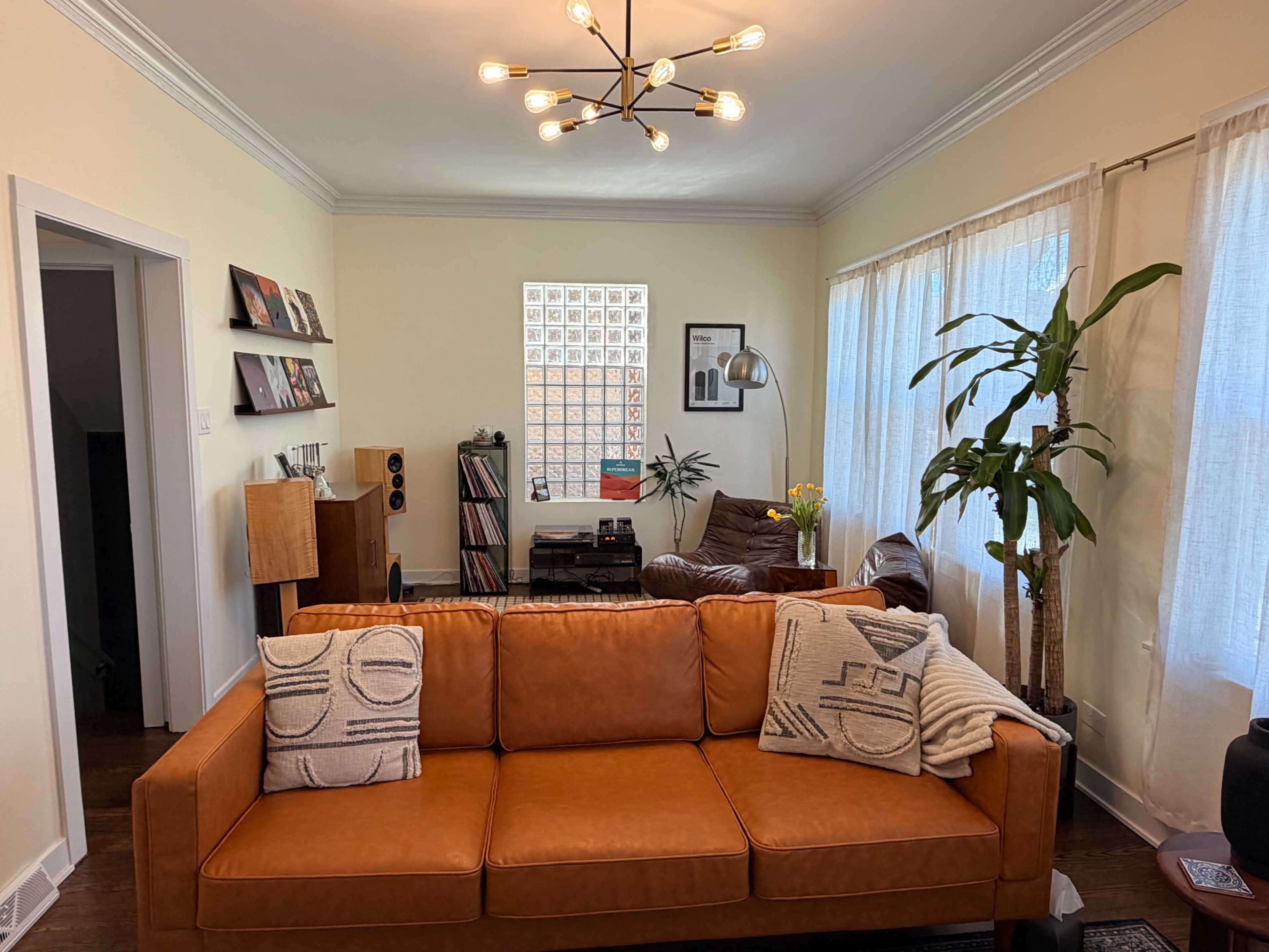 A modern living room features a brown leather sofa, a record player, and large windows with sheer curtains, creating a cozy atmosphere.