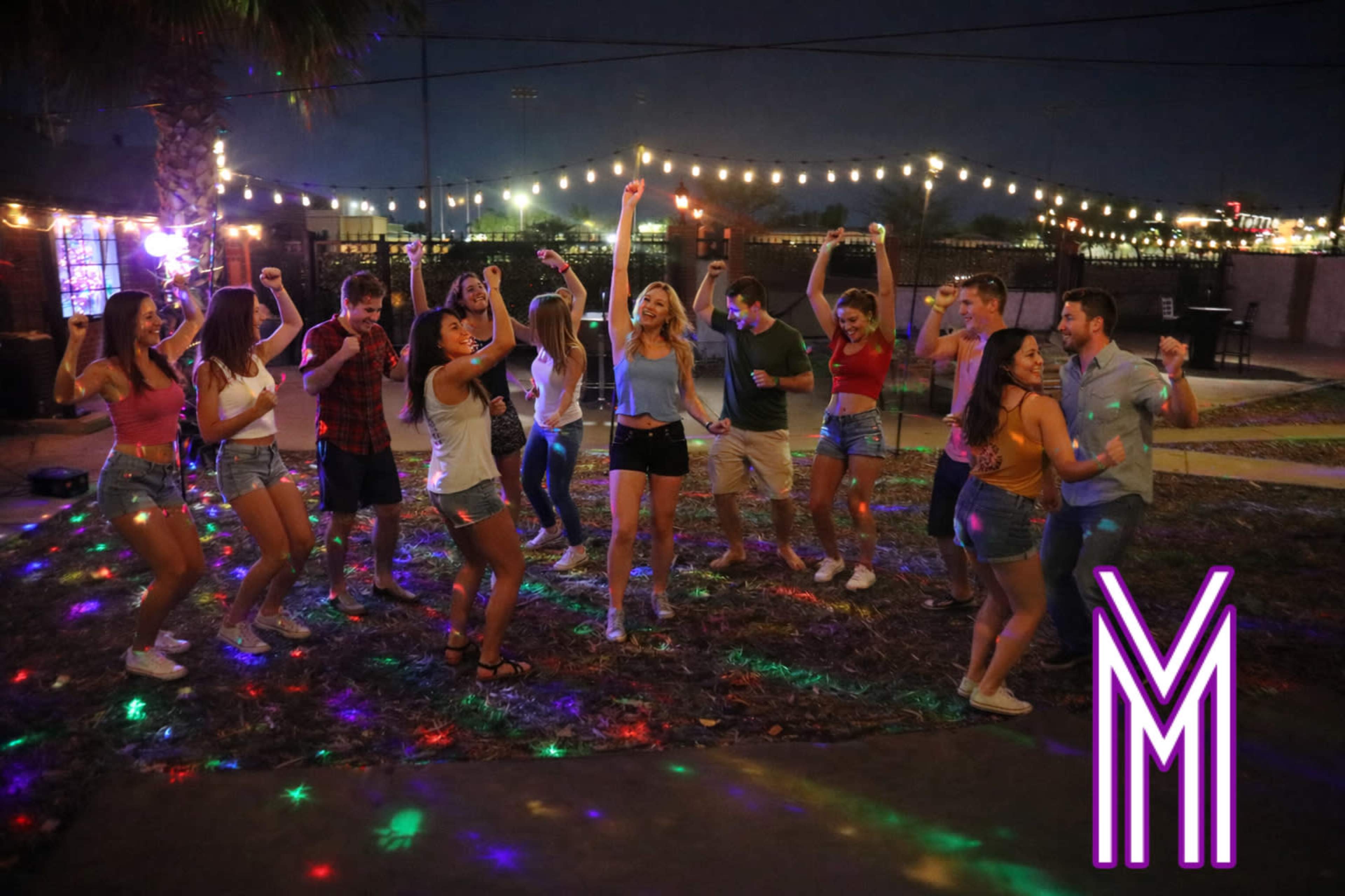 A group of young adults is dancing outdoors under string lights and colorful disco lights during a nighttime celebration.