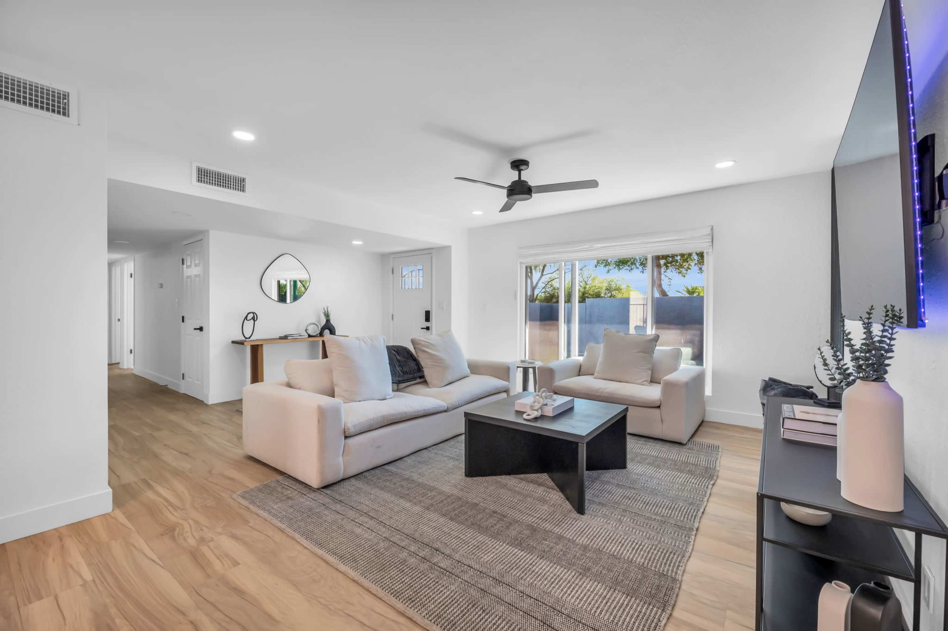 The room features a modern living area with a light-colored sofa set, a dark coffee table, and large windows that let in natural light.