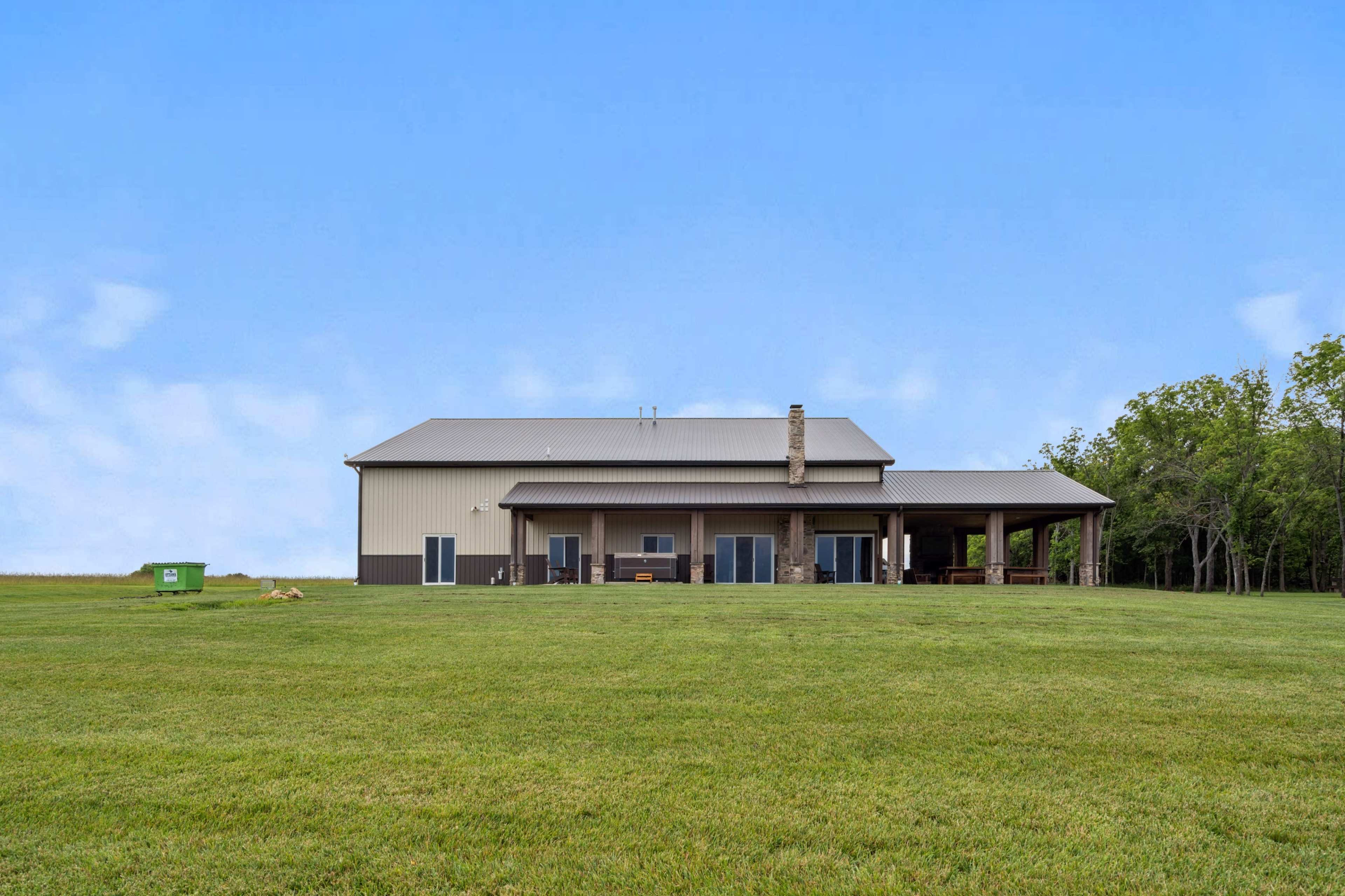A large modern house with a metal exterior sits on a grassy expanse, with a covered porch and several glass doors facing the yard.