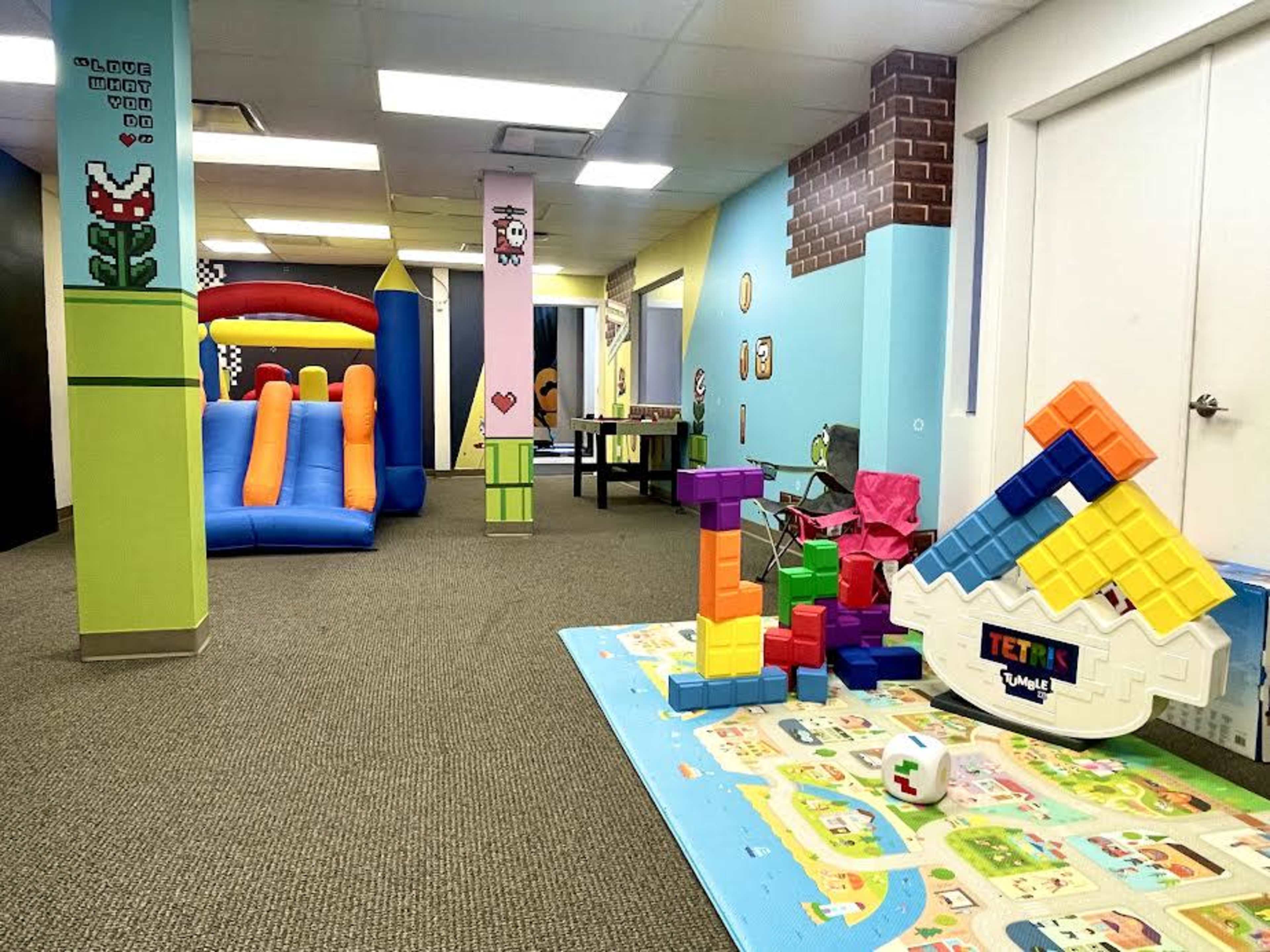 The image shows a colorful indoor play area featuring an inflatable slide, a table with toys, and a large puzzle mat on the floor.